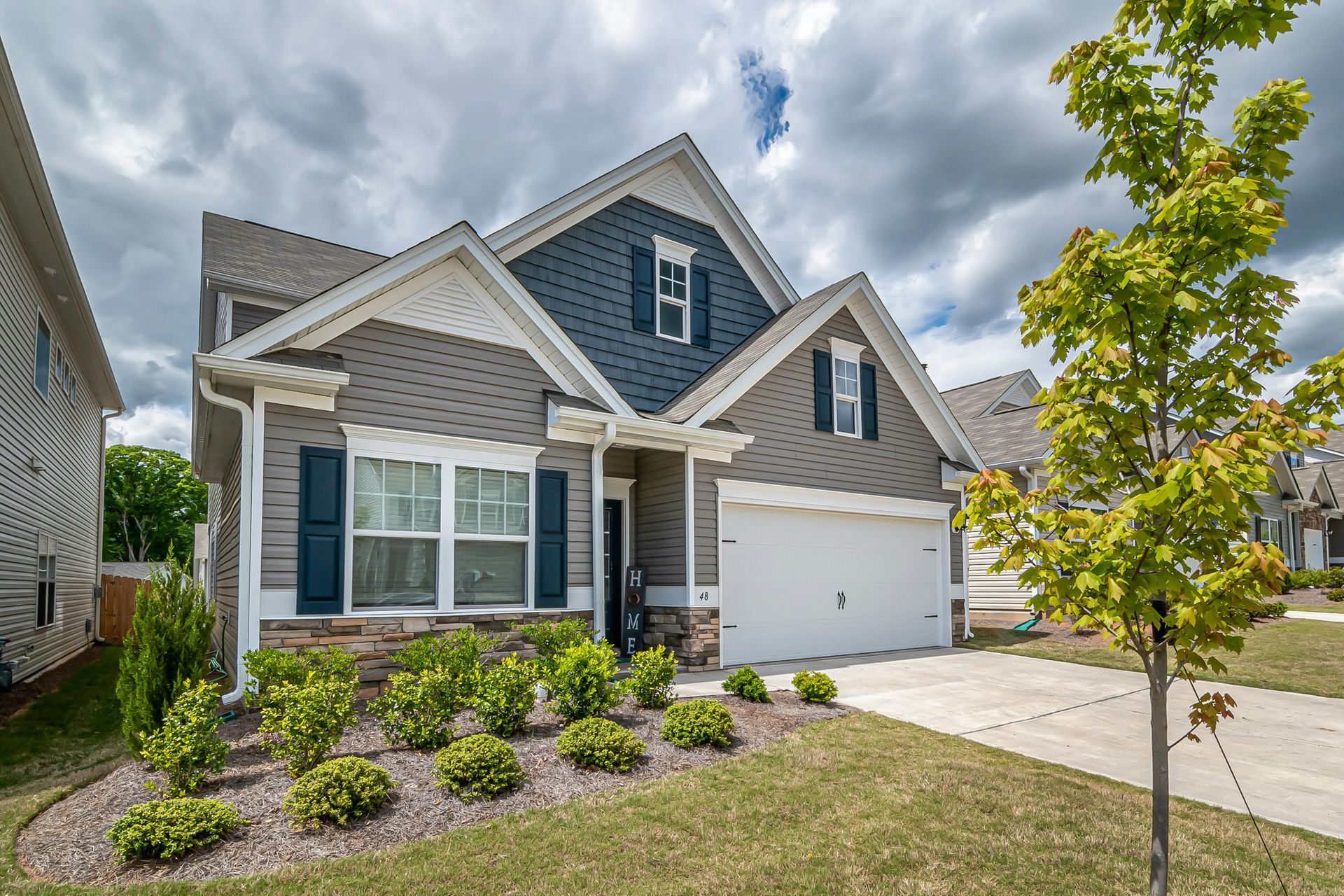 A two-story suburban house with gray siding, blue accents, a white garage, and a landscaped front yard under a cloudy sky.