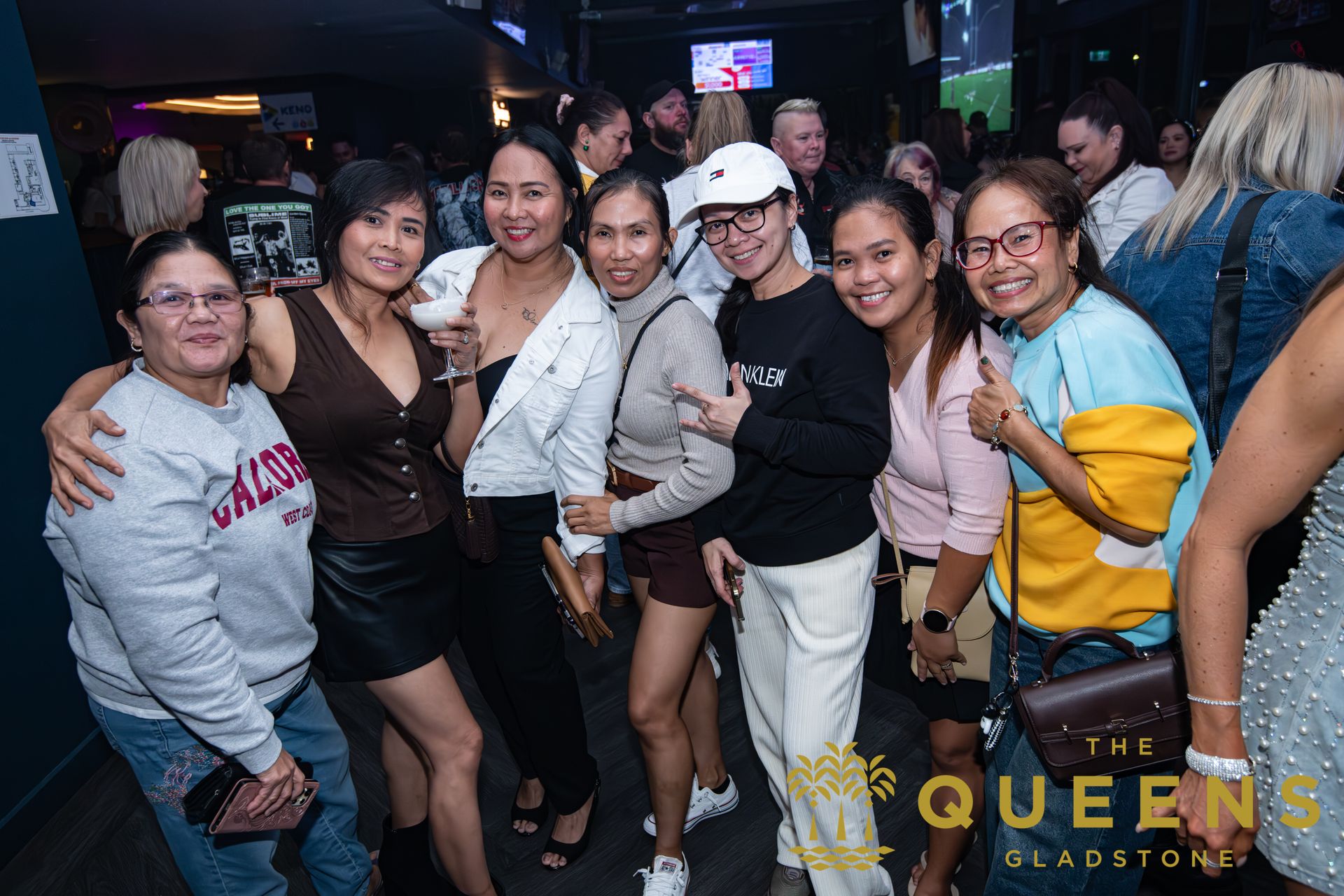 Group of smiling friends posing together in a dimly lit nightclub, one holding a drink.