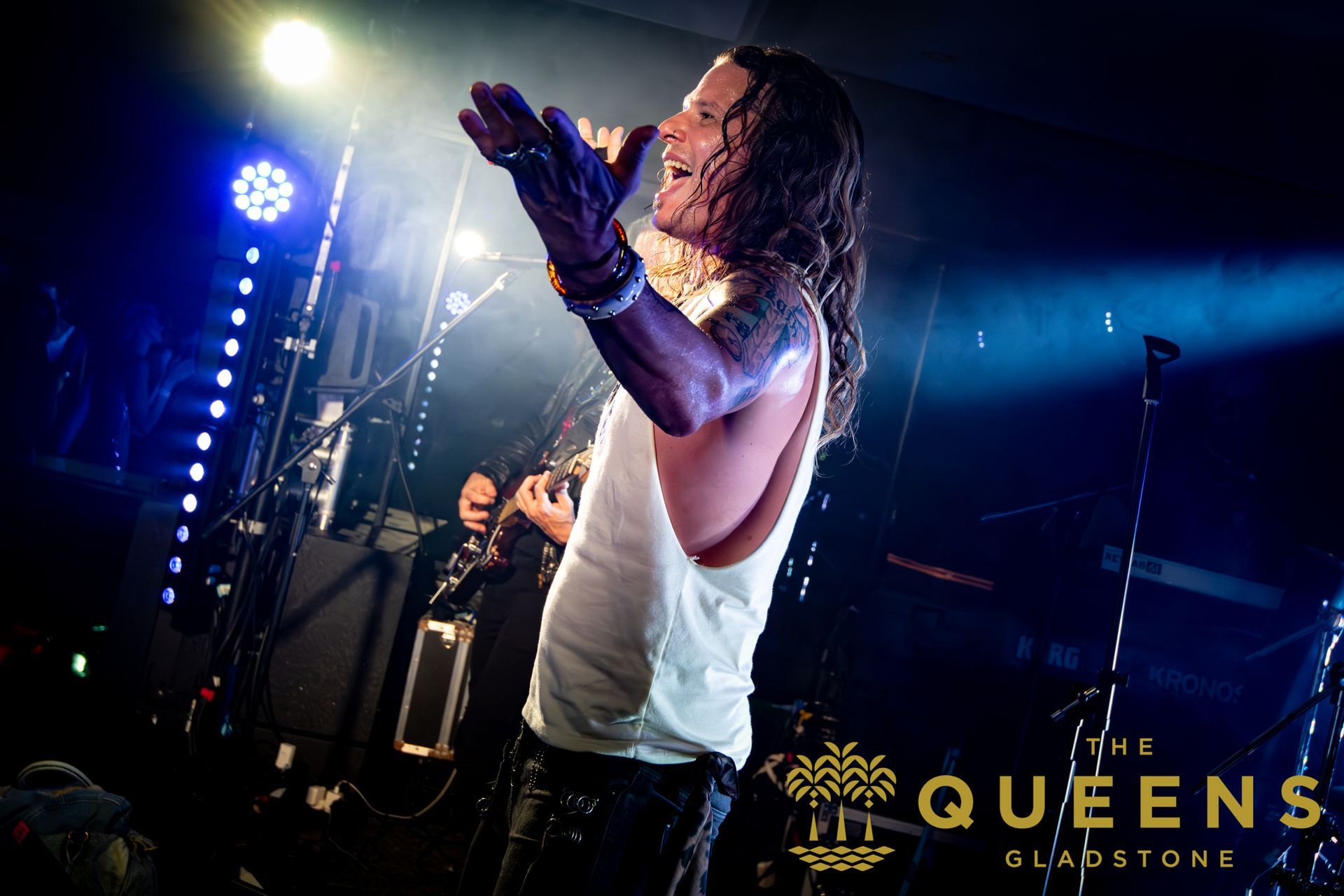 Singer in white tank top performing onstage under blue lights, with “The Queens” logo in the corner