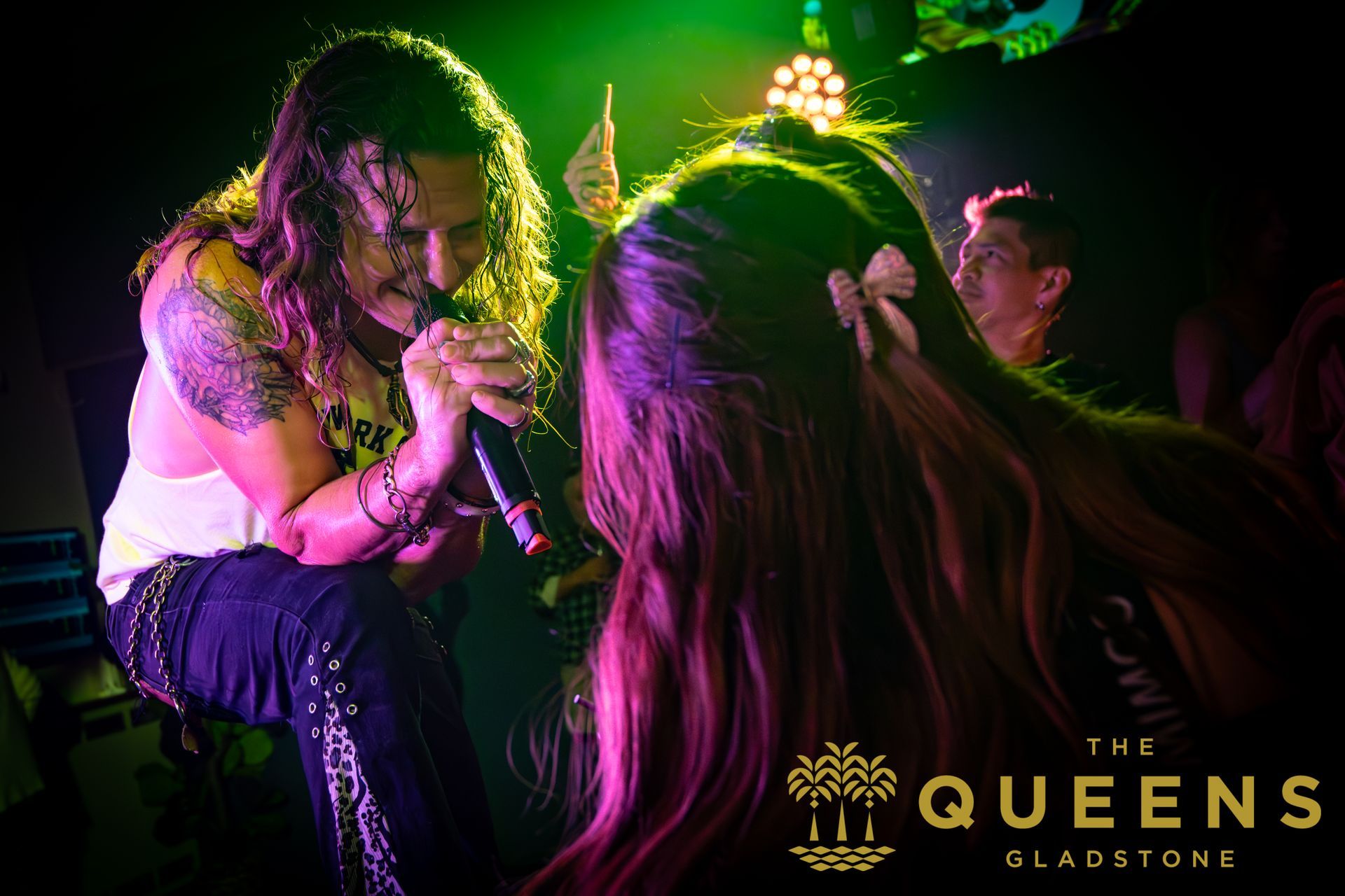 Two performers under green and pink lights, one crouched singing to the other, with “The Queens Glasgow” text.
