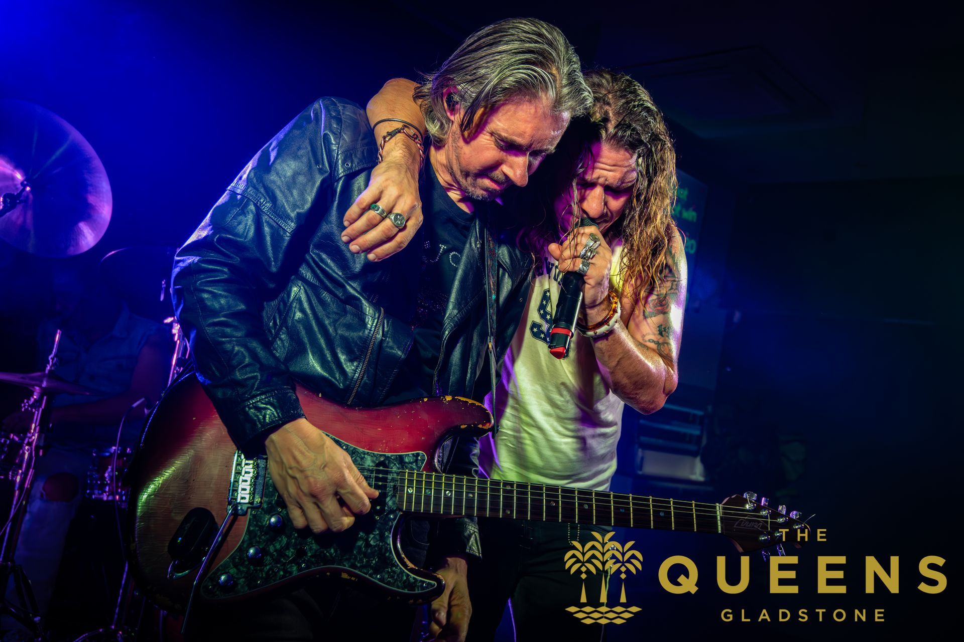 Two musicians onstage, one hugging the other while playing guitar, under purple-blue lights with “Queens” logo.