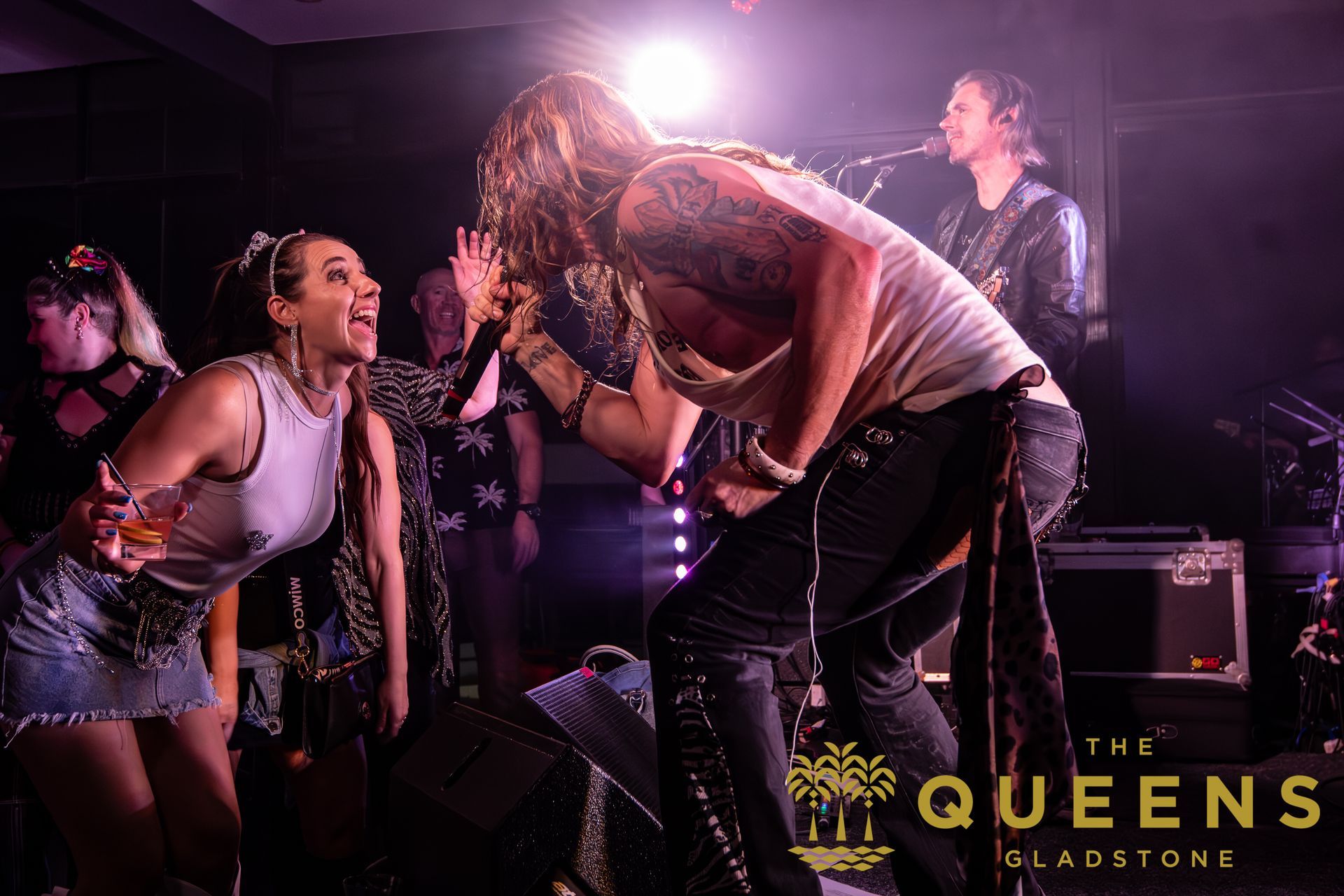 Live band performance with singer bent over singing to an excited crowd under pink stage lights.