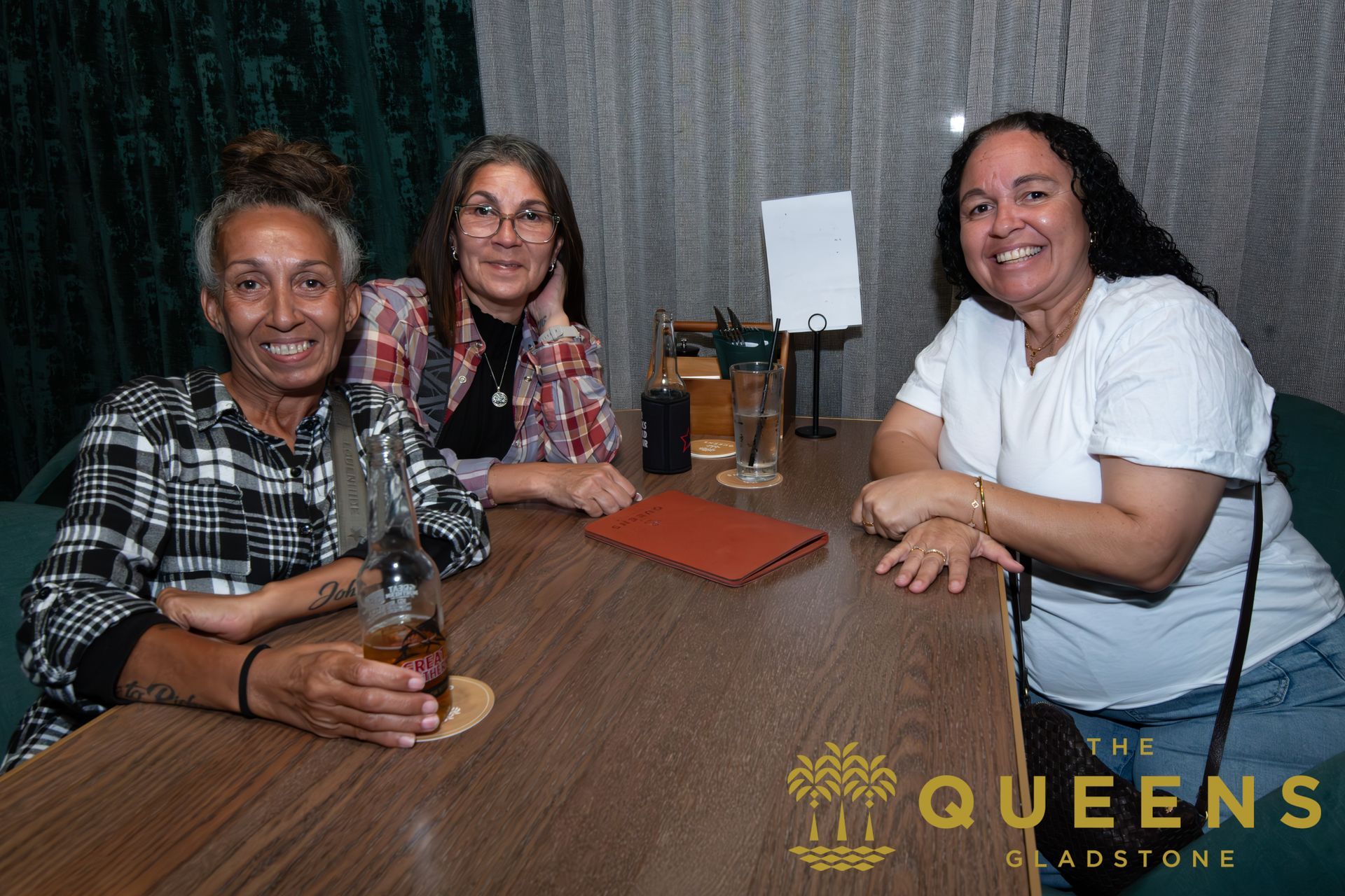 Three people smiling at a restaurant table with drinks and menus, Queen’s Roadhouse logo visible