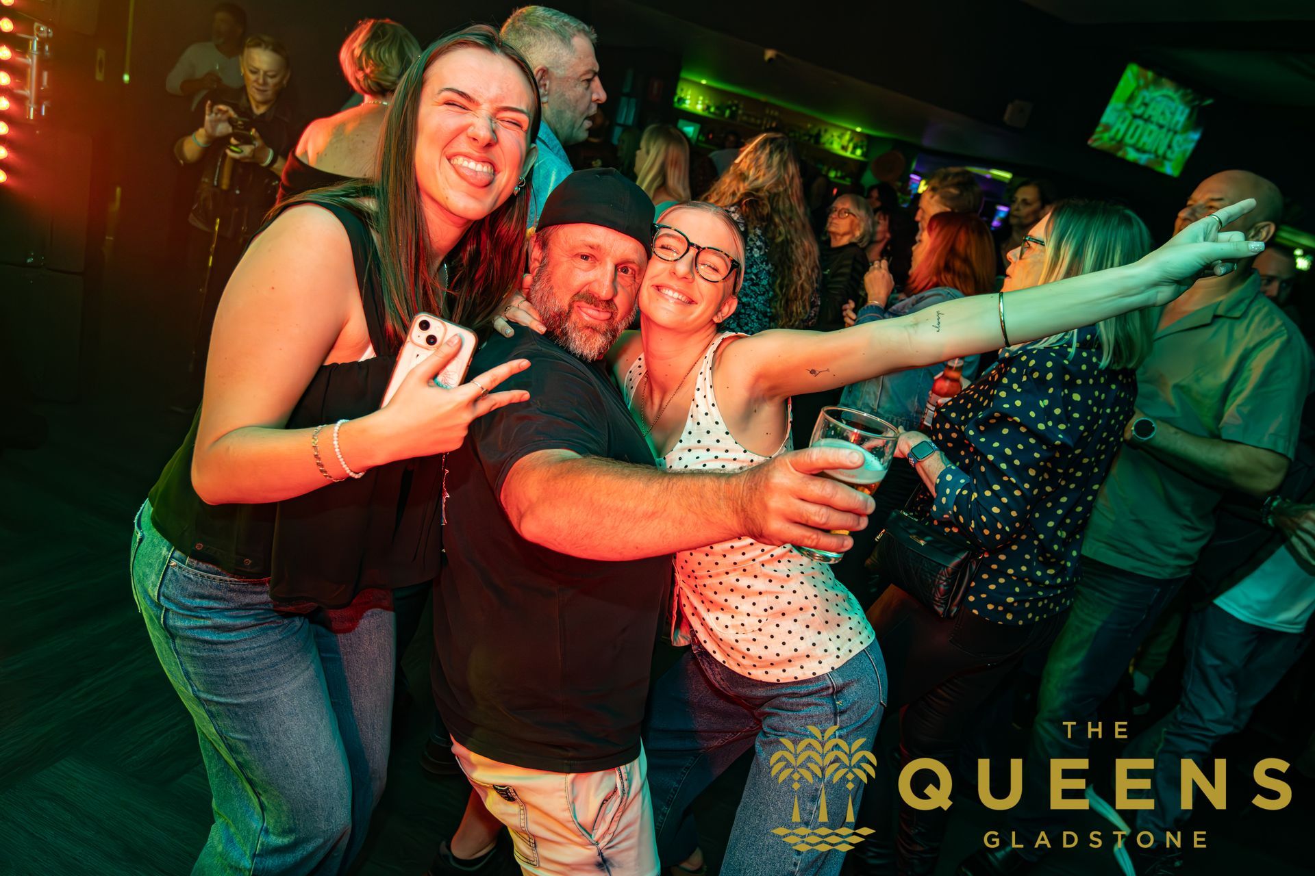 Smiling group posing in a lively club, with green lighting and a “The Queens” sign in the background