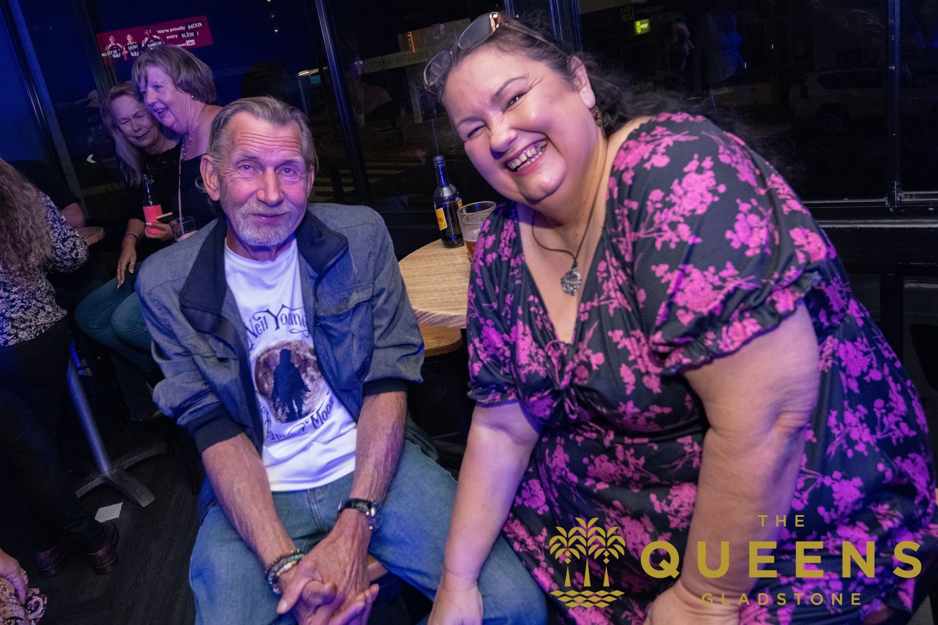 Two people smiling and posing at a nightclub, with colorful lighting and the word “QUEENS” visible.