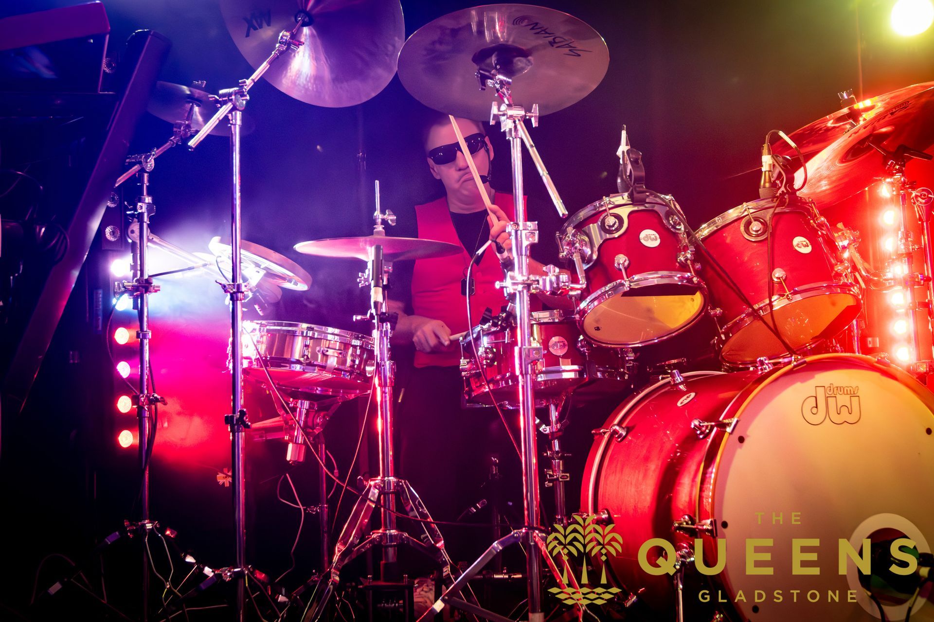 Drummer on a red Queen’s stage under pink-purple lights, playing behind a drum kit.