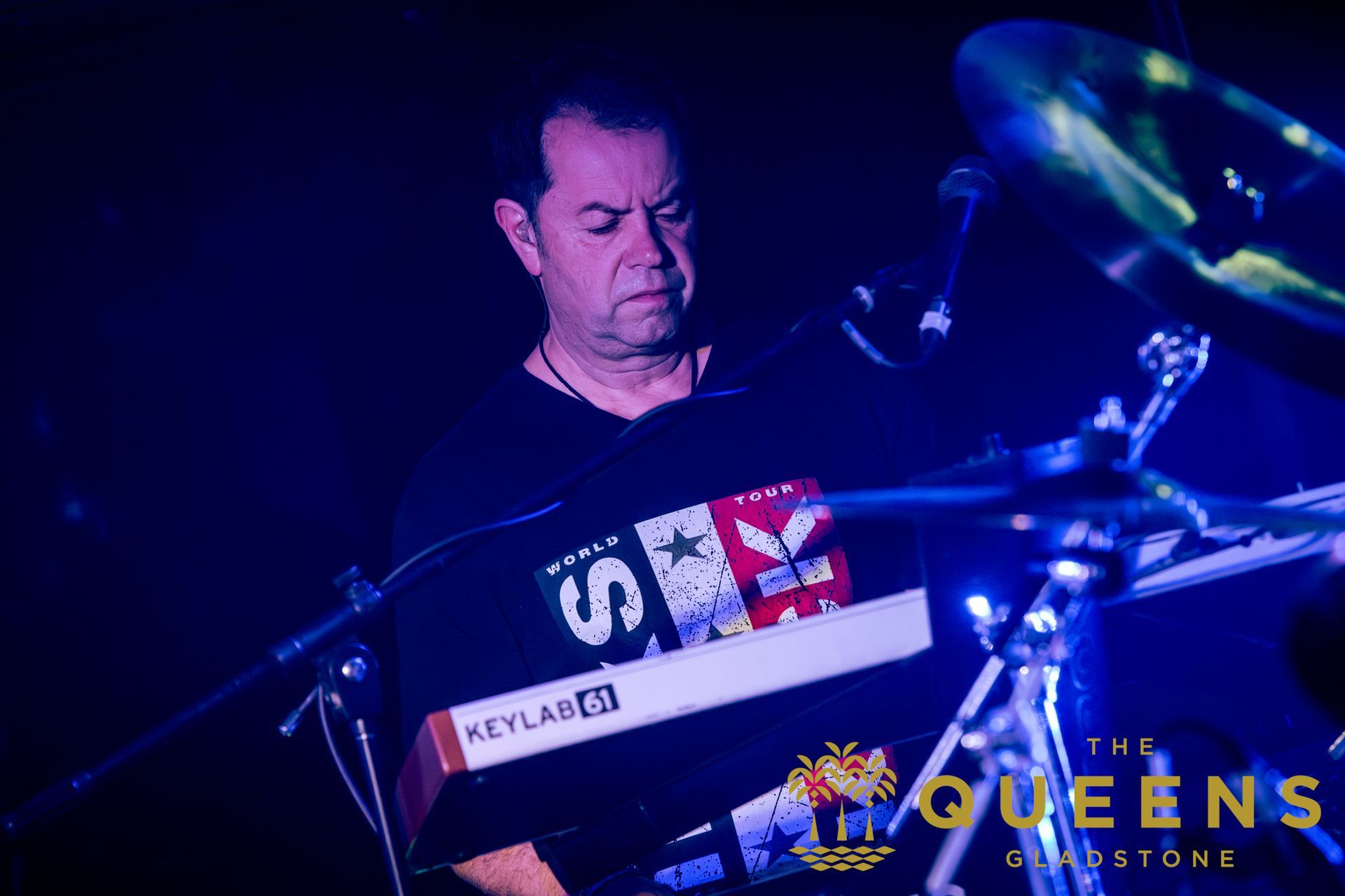 Drummer playing on stage under blue lights, with a Queen-themed setup visible on the drum kit.