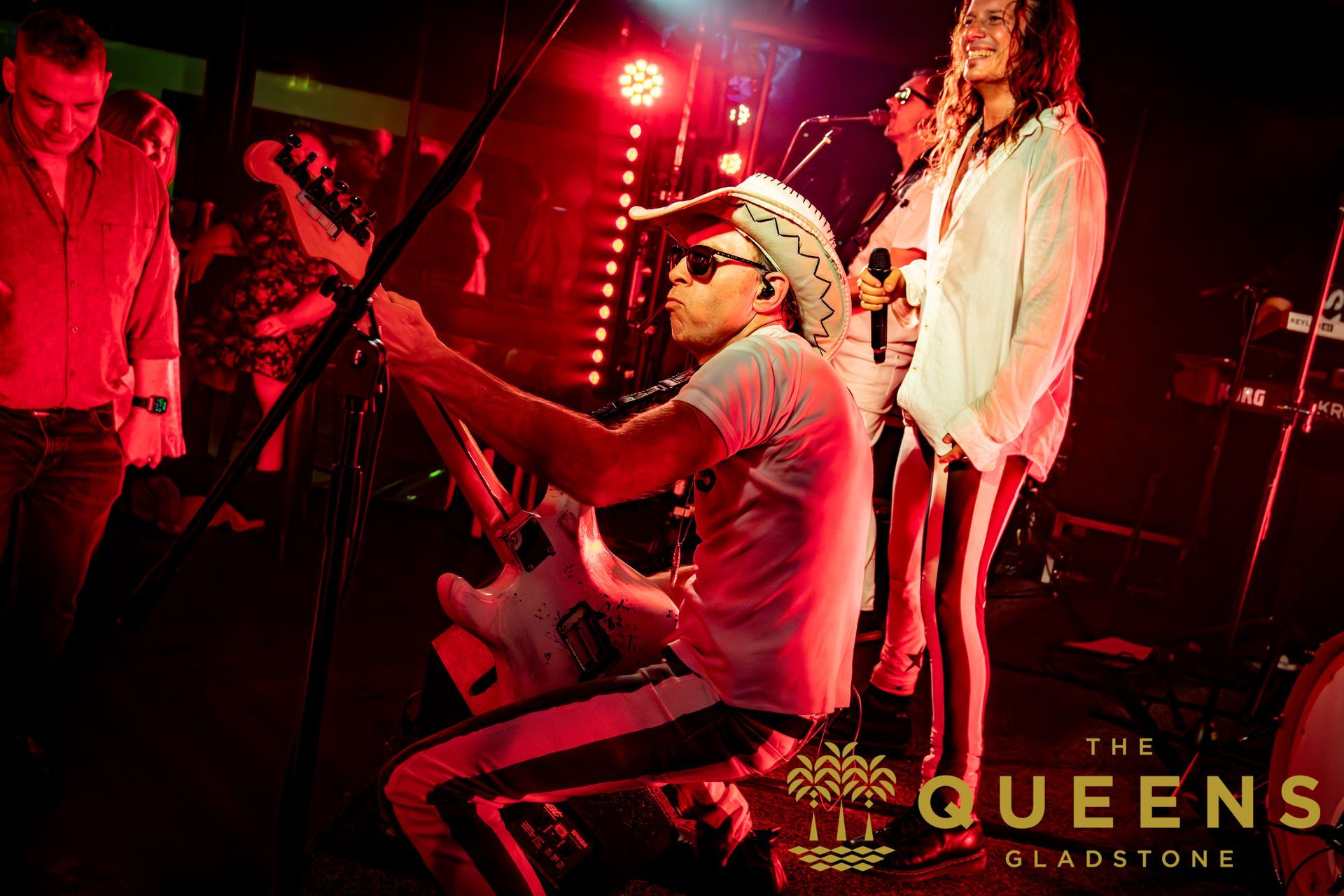Concert stage with red lighting, guitarist seated center, singer at right, and “The Queens” text
