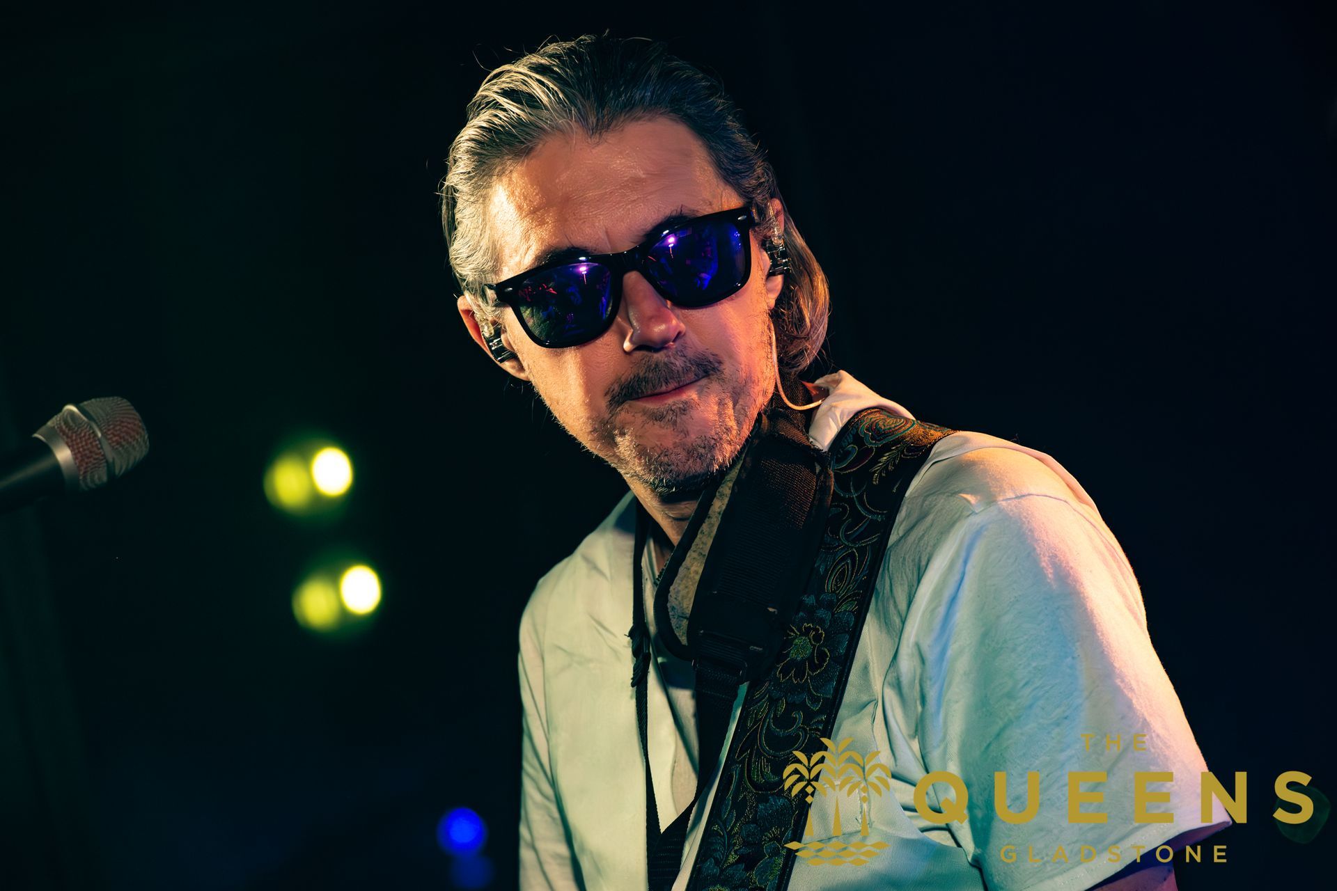 Musician wearing sunglasses and a patterned jacket onstage under green and blue lights