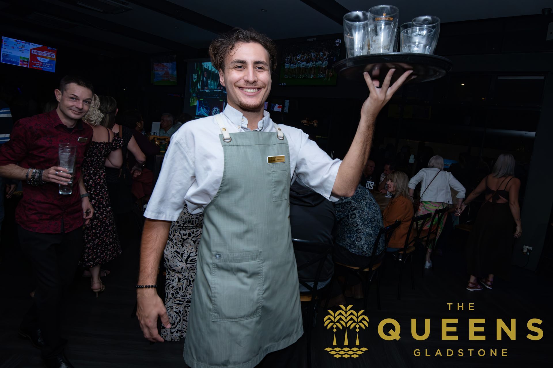 Waiter balancing a tray of glasses in a dimly lit bar, with “The Queens Gladstone” sign visible.
