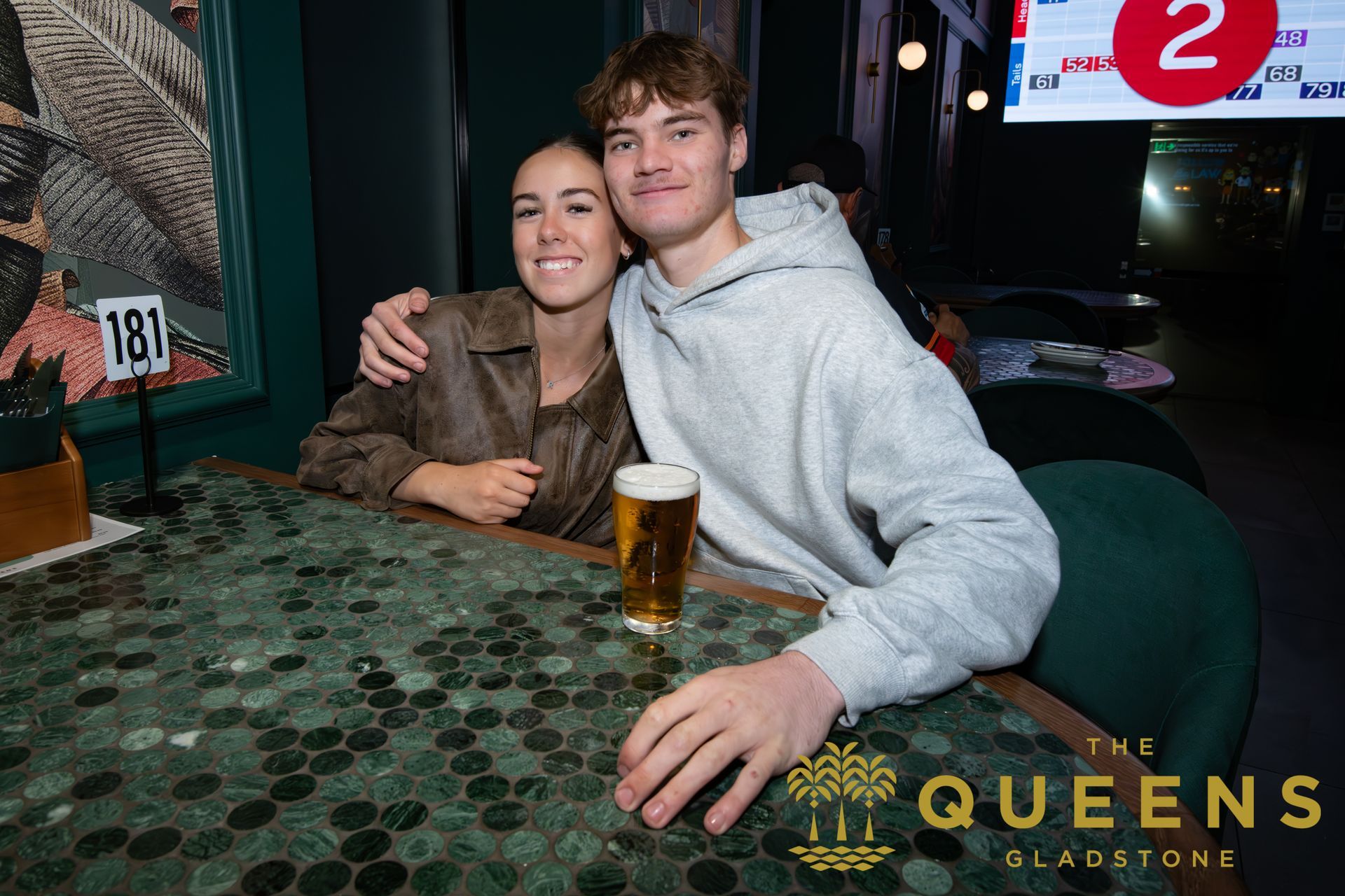 Smiling couple seated at a café table with a pint of beer, QUEENS sign in the background