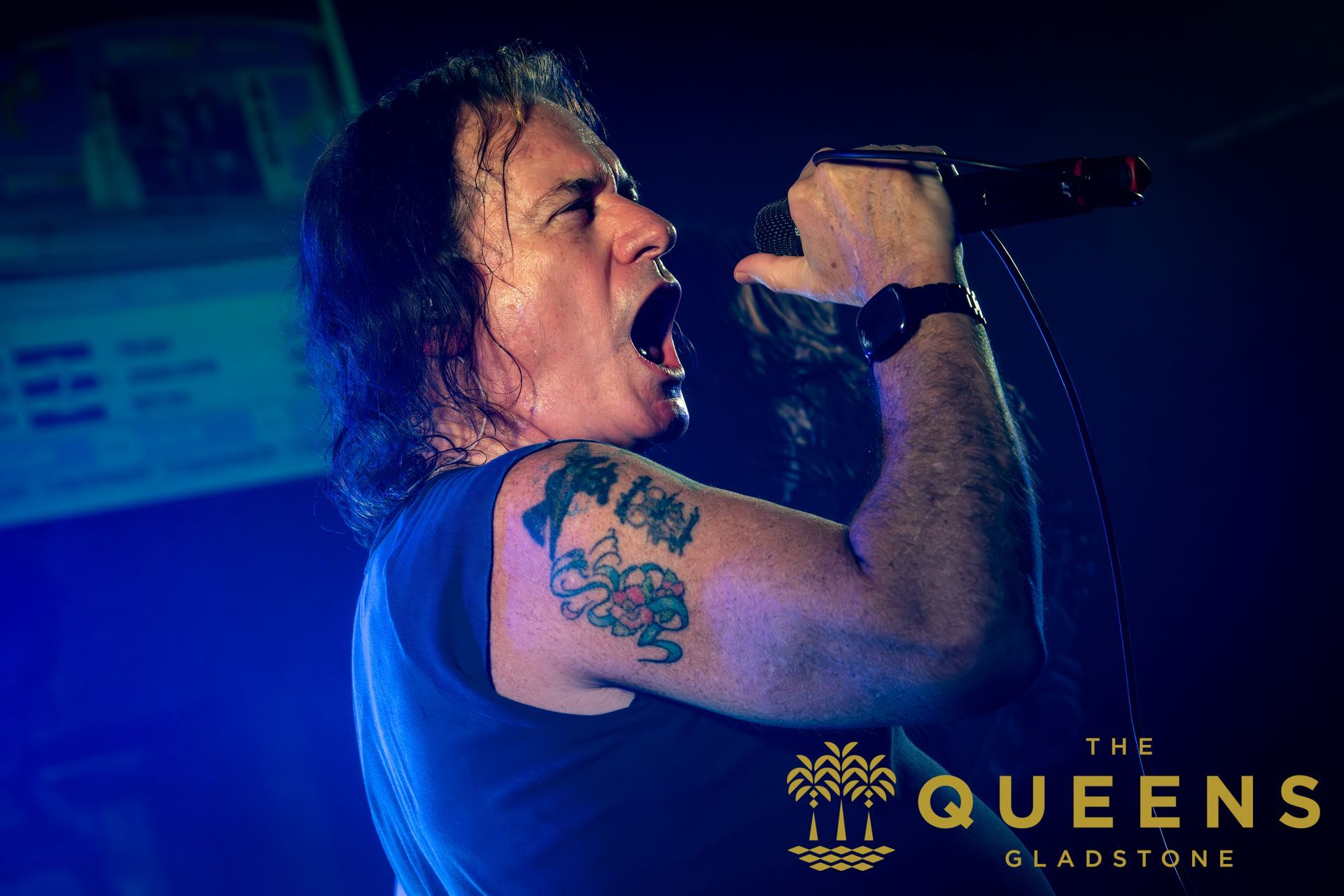 Singer shouting into a microphone on a blue-lit stage, with a tattooed arm and “The Queens” logo.