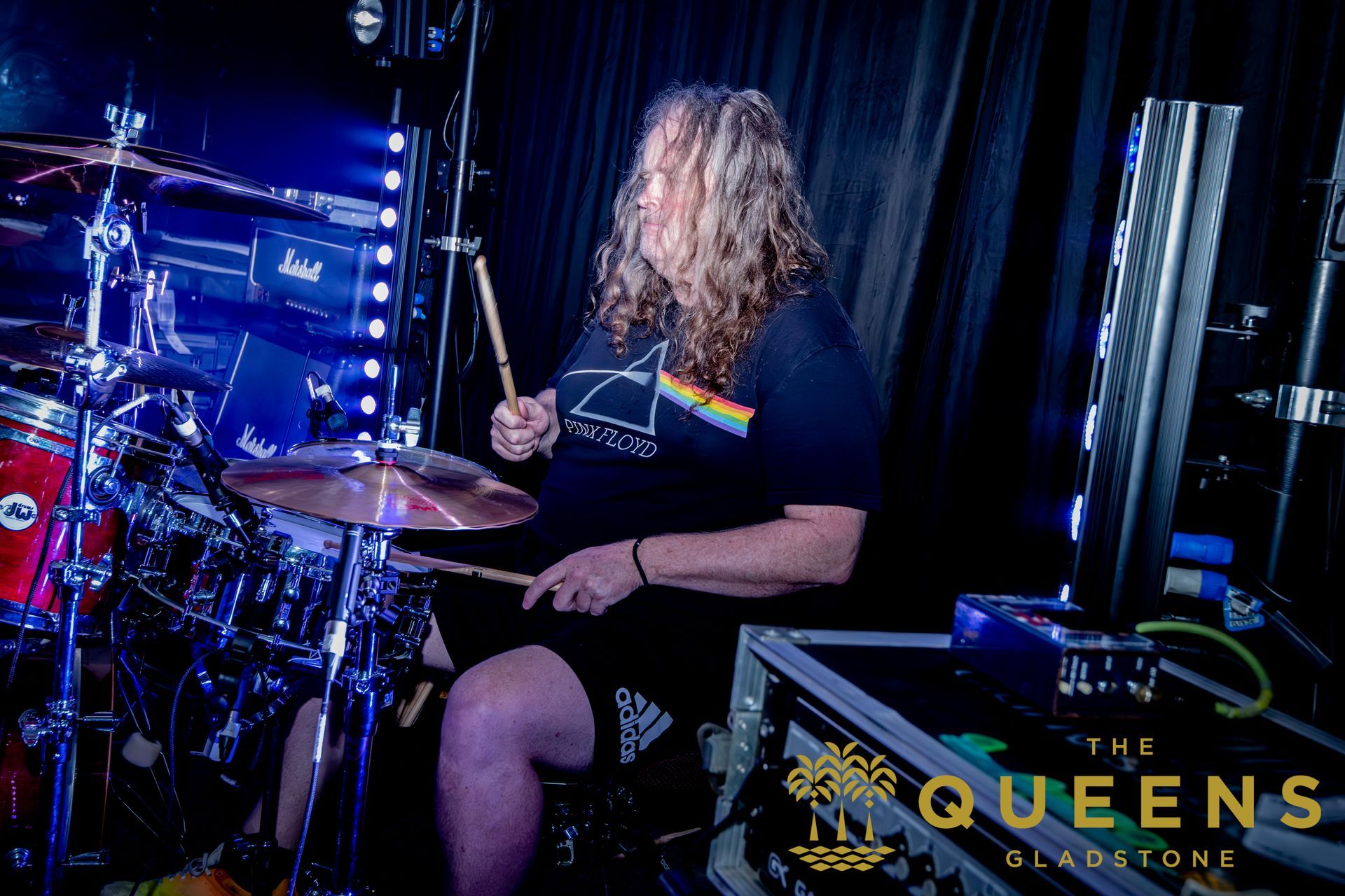 Drummer onstage under blue lights, playing a drum kit with “Queens” logo in the corner.