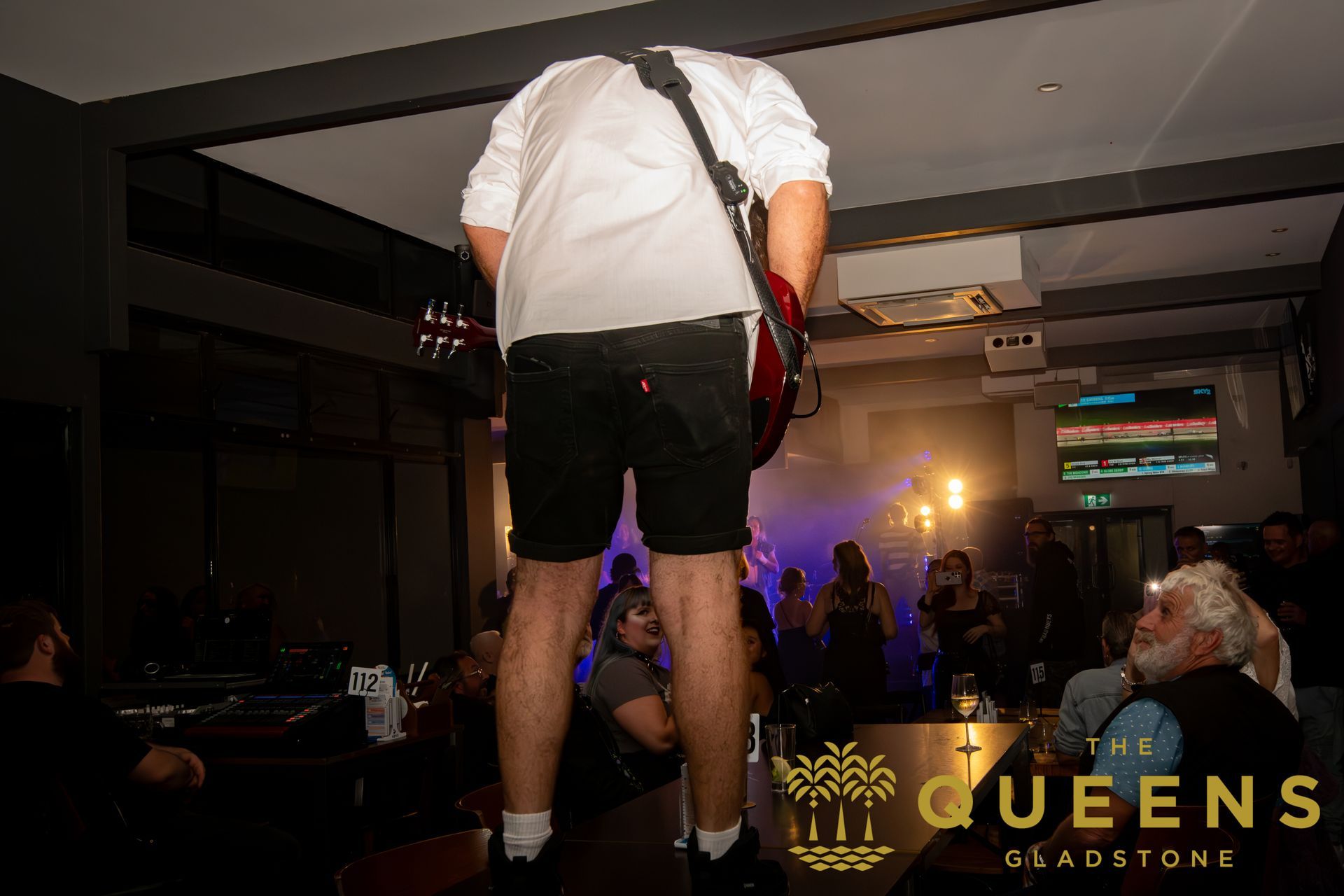 Musician onstage in a white shirt and black shorts, facing a dim crowd under purple lights at Queens Ballroom.