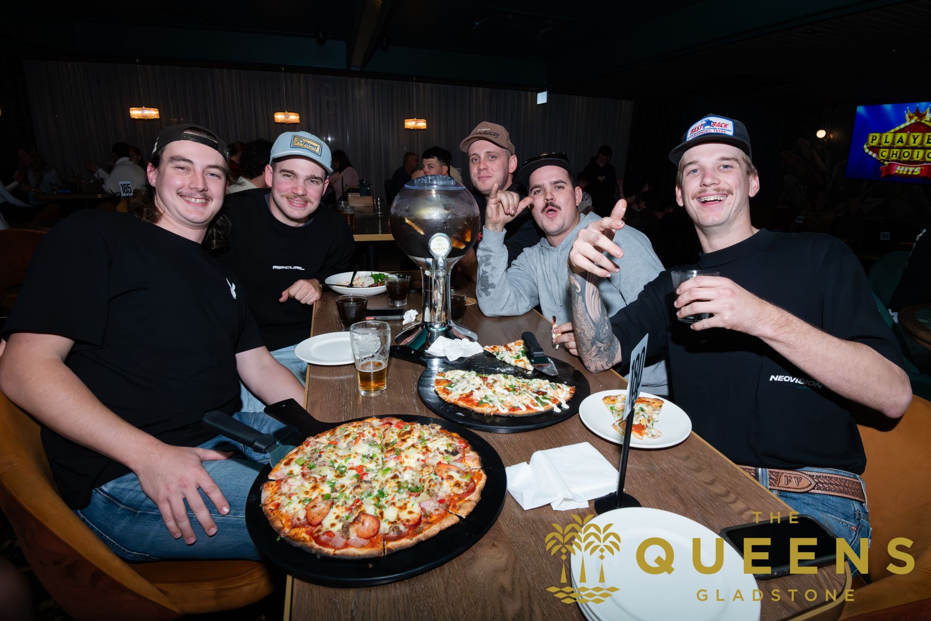 Group of friends at a restaurant table with pizza and drinks, smiling and toasting around a trophy-like centerpiece.