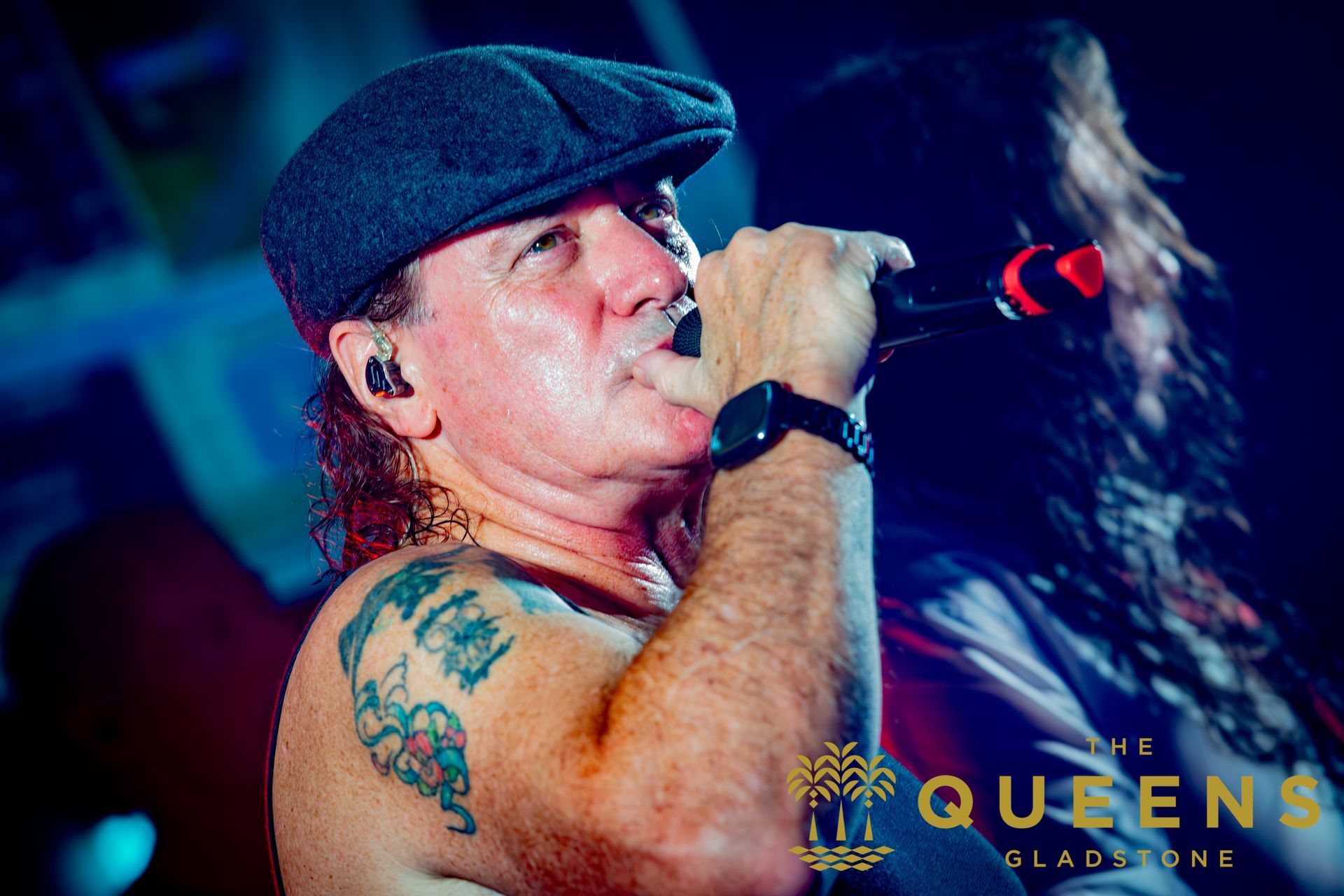 Singer in a flat cap performs into a microphone under blue stage lights, with a tattoo visible on his arm.