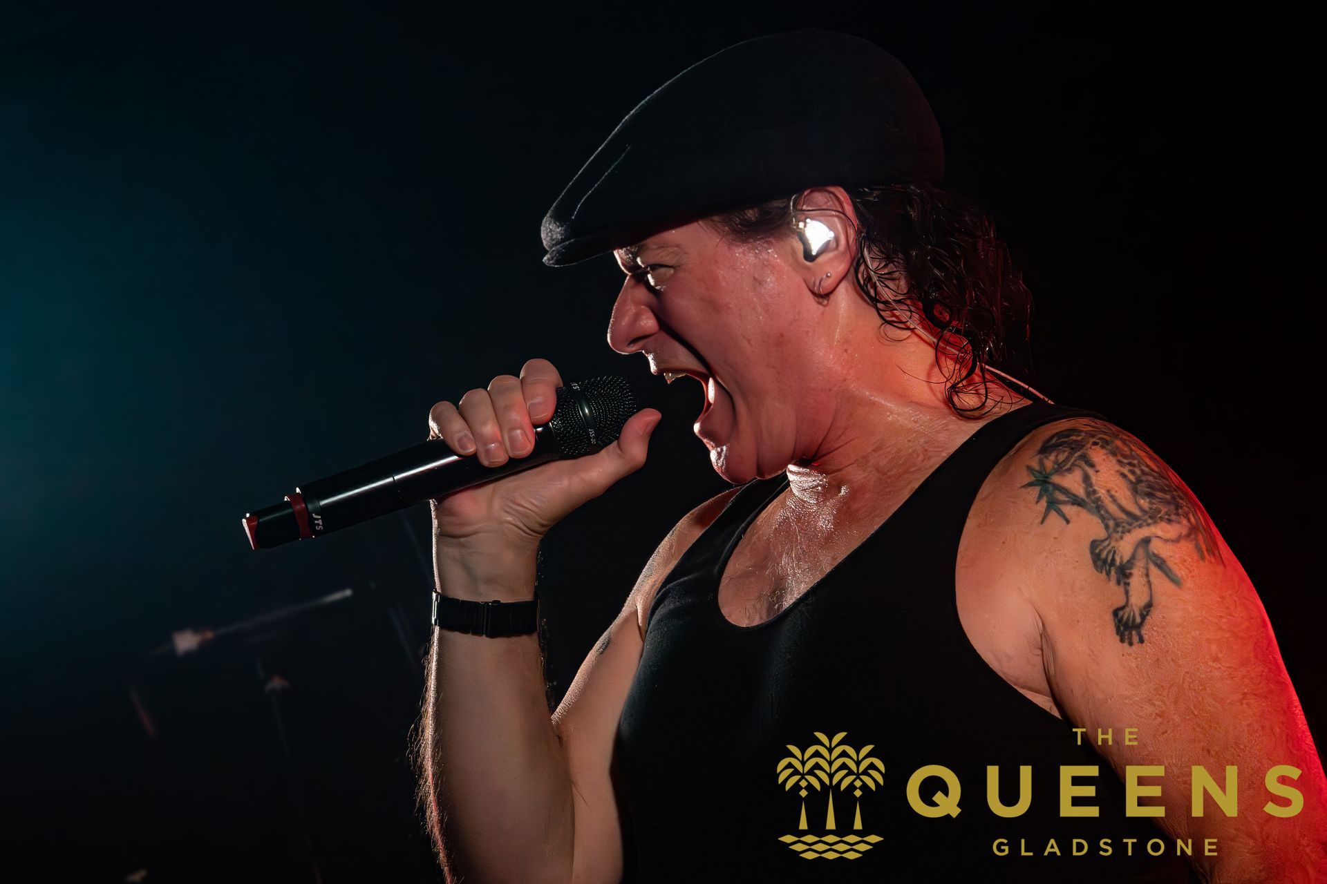 Singer in black cap and tank top singing into a microphone on a dark stage, with “Queens” text at bottom right