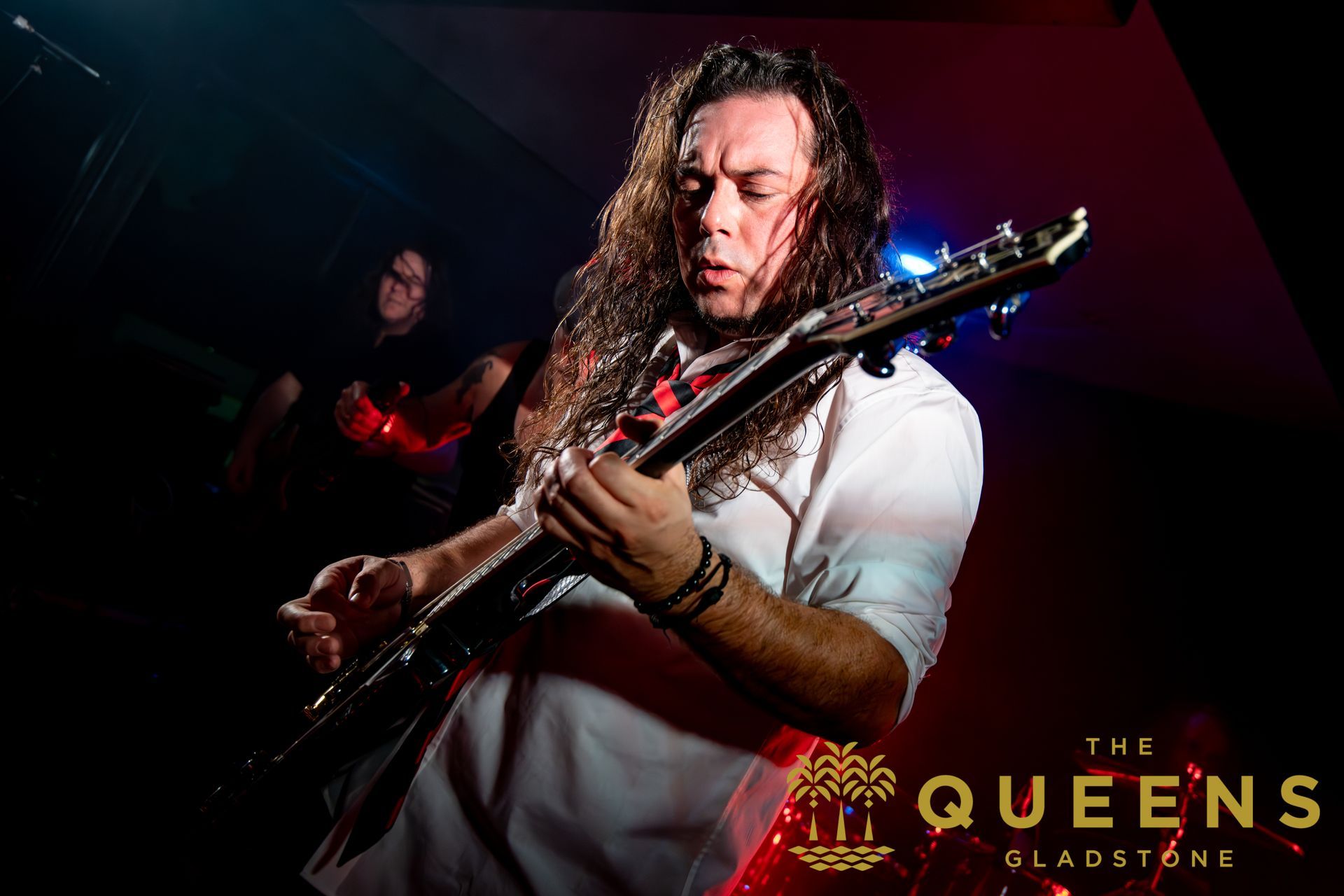 Guitarist performing under red and blue stage lights, with “The Queens of the Stone Age” text in the corner