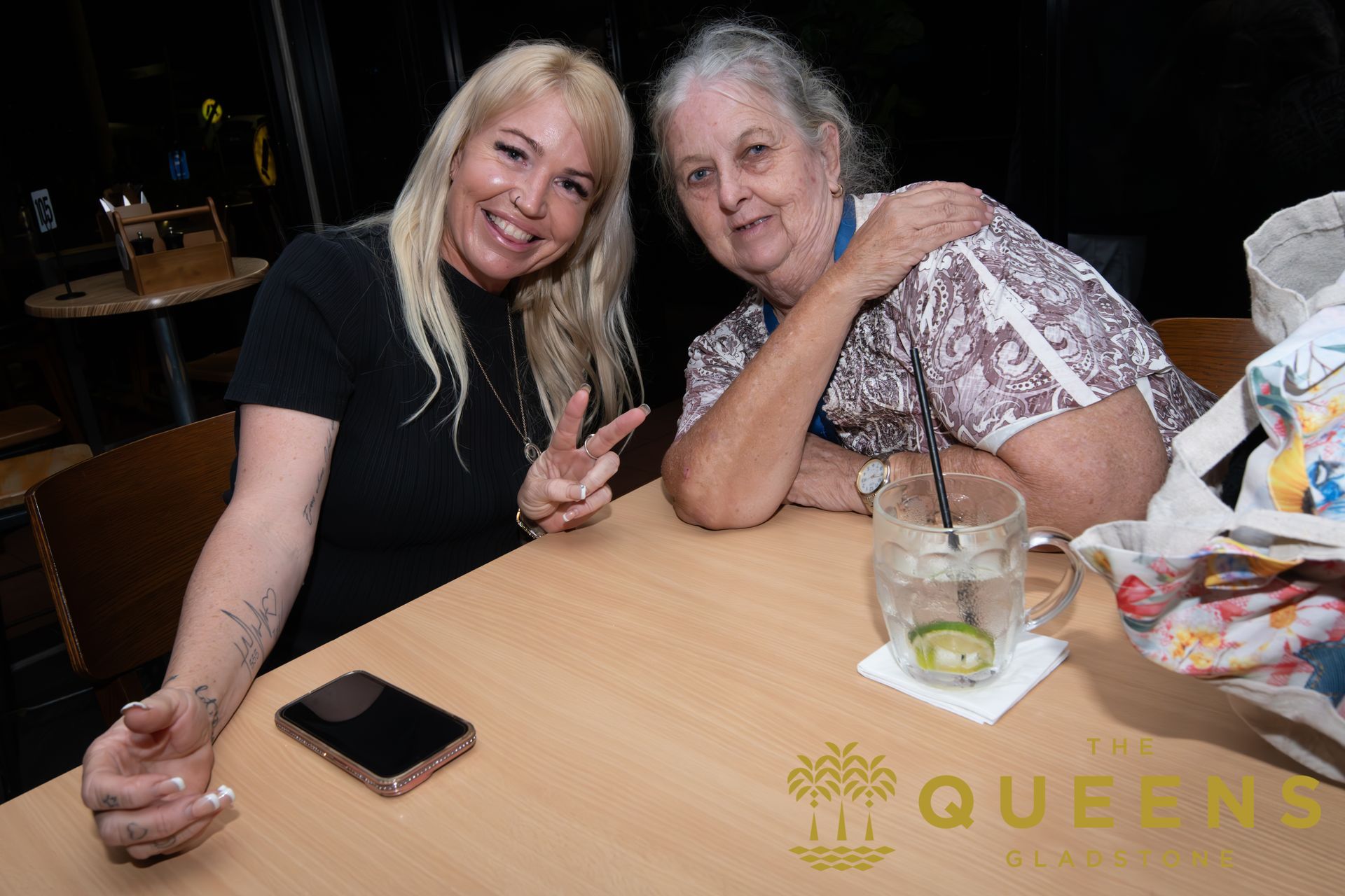 Two women smiling at a table, posing with a peace sign, with drinks and a phone in front of them.