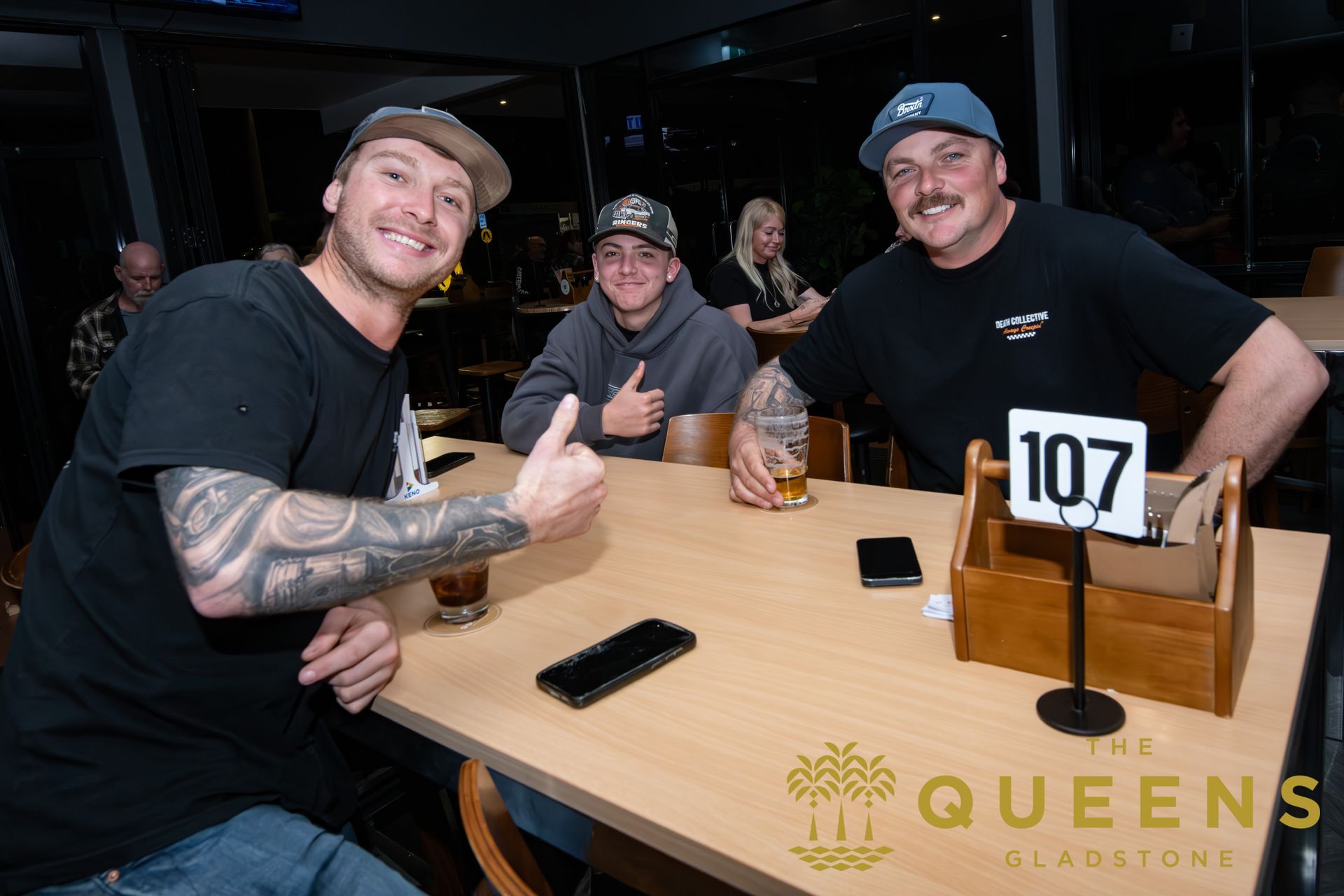 Three men smiling and giving thumbs-up at a table in a restaurant, with a table number 107 sign.