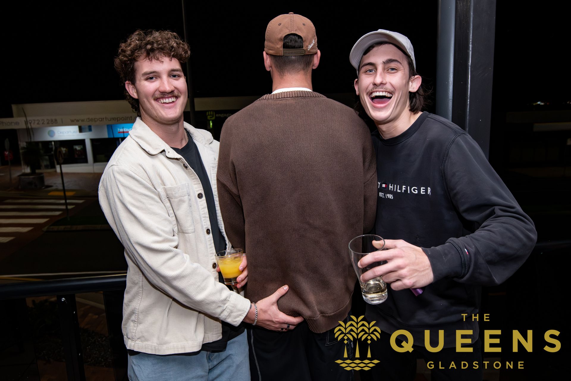 Three people posing at a bar, one turning away, with a “The Queens Gladstone” logo in the corner.