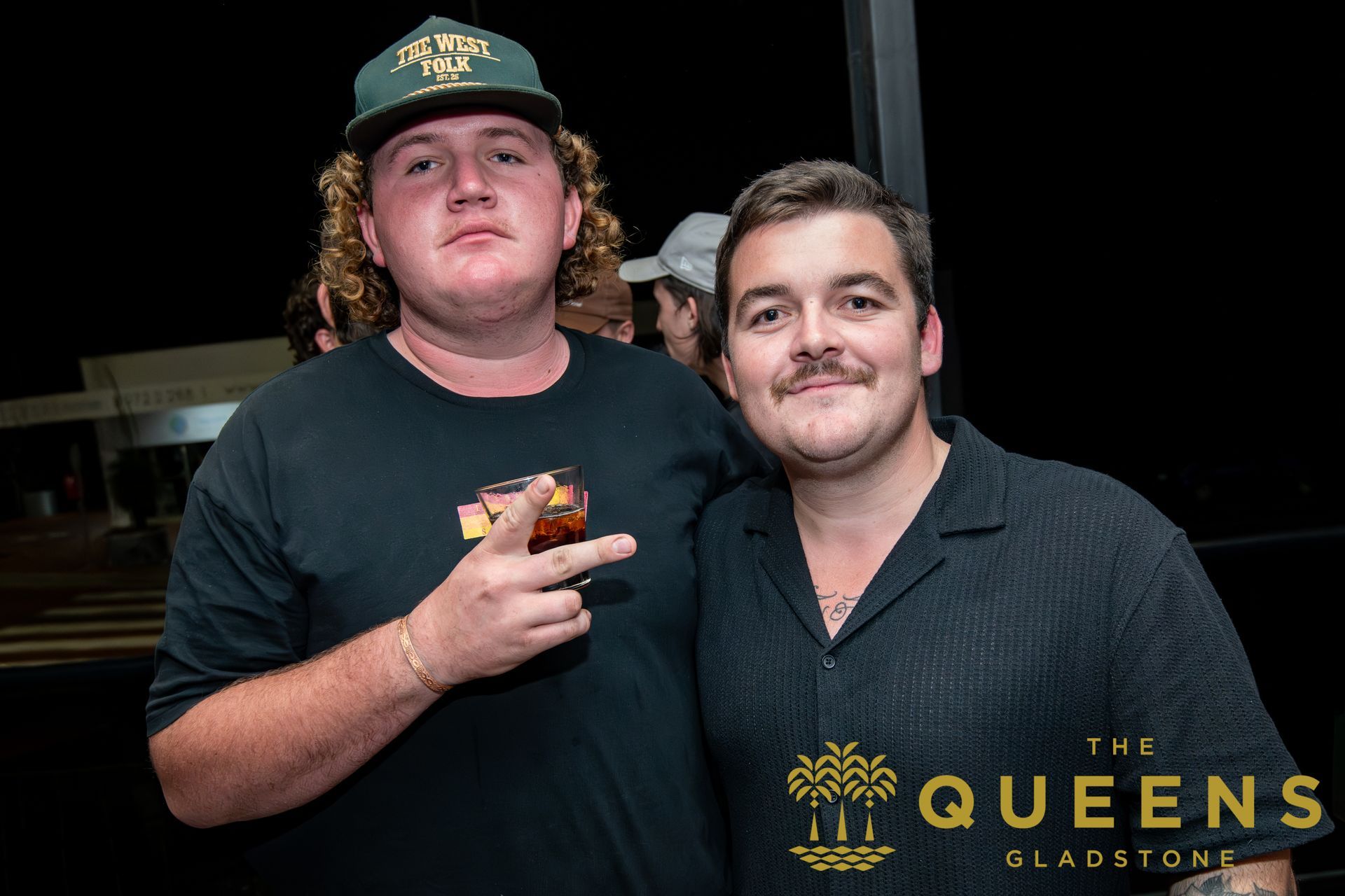 Two men posing at a nightclub, one holding a drink, with The Queens Sladston sign in the background