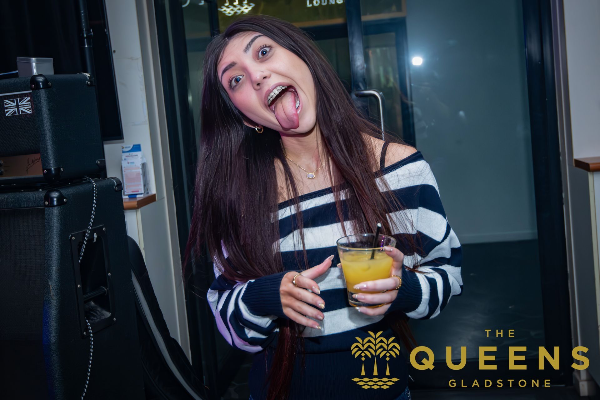 Woman at a bar smiling and holding a yellow drink, with the Queen's Sladstone logo in the corner