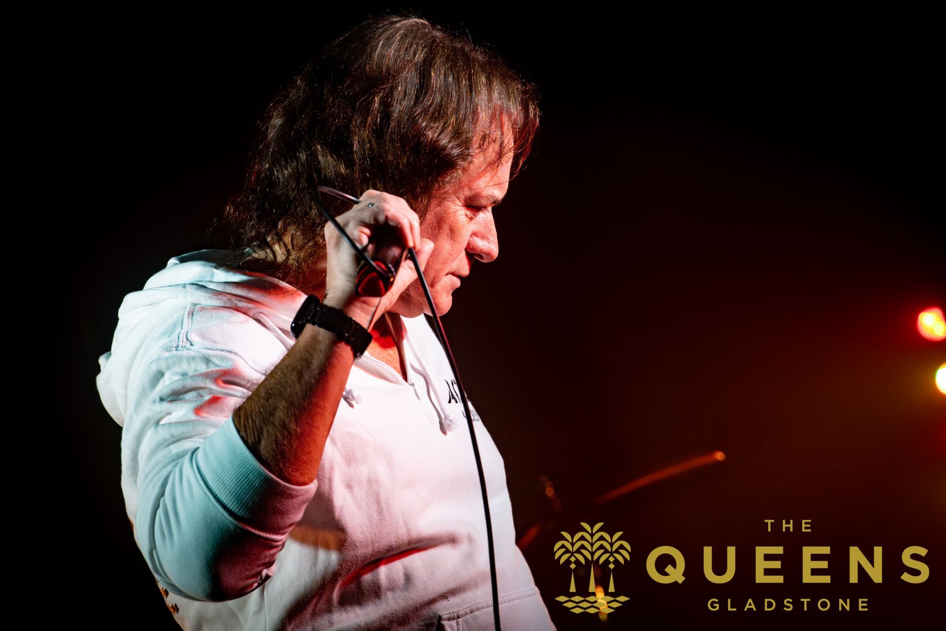 Singer in white shirt holding a microphone on a dark stage, with The Queens Gladstone logo visible