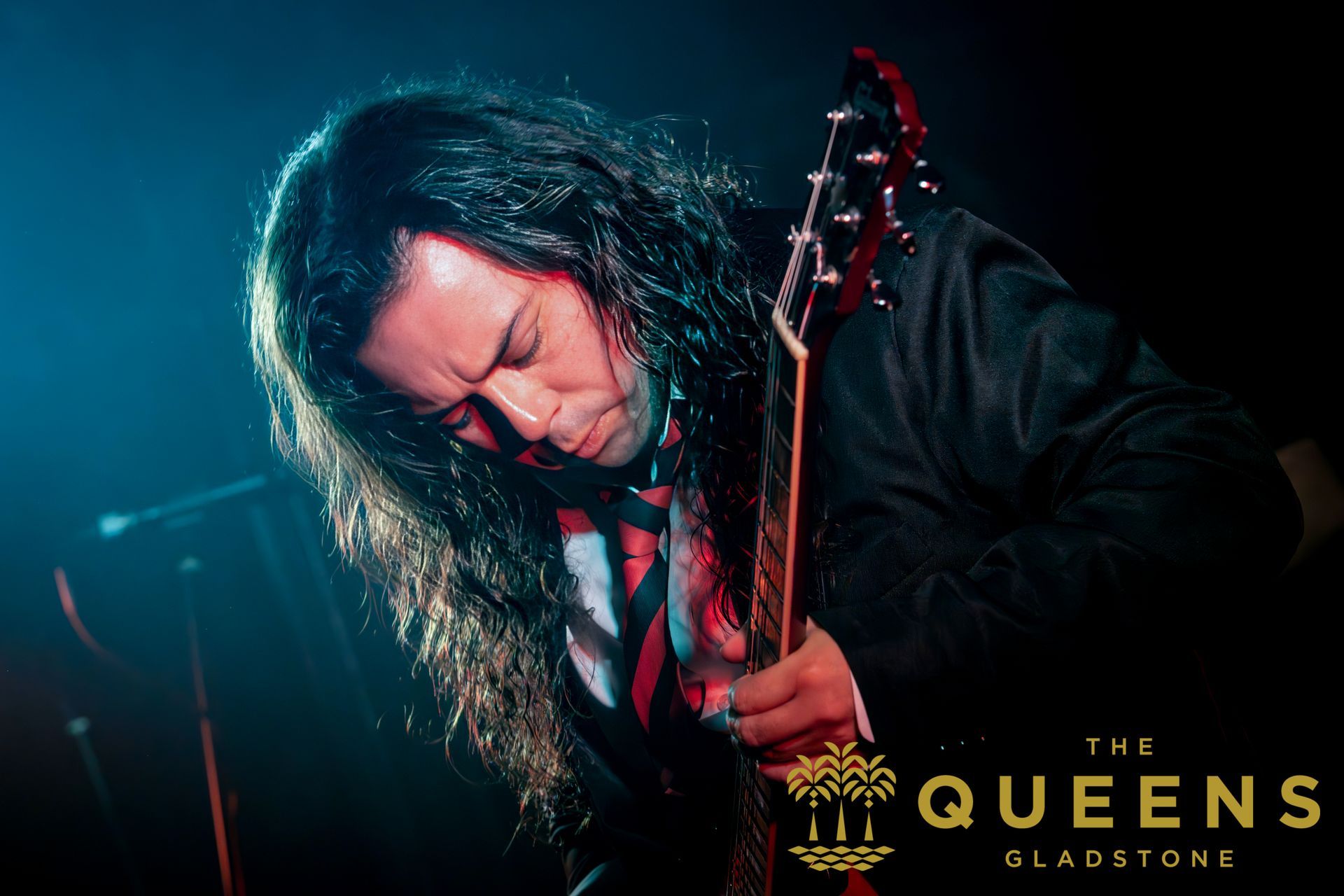Guitarist on a dark stage with red-blue lighting, head bowed while playing, “The Queens” logo in the corner