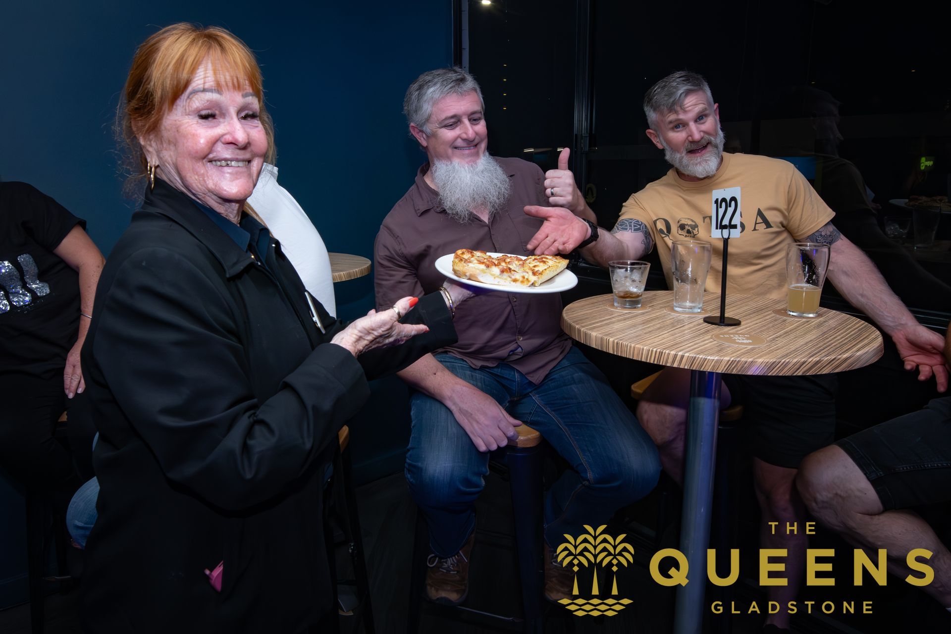 Three people at a nightclub table; one woman smiles while holding a drink, with The Queens logo visible.