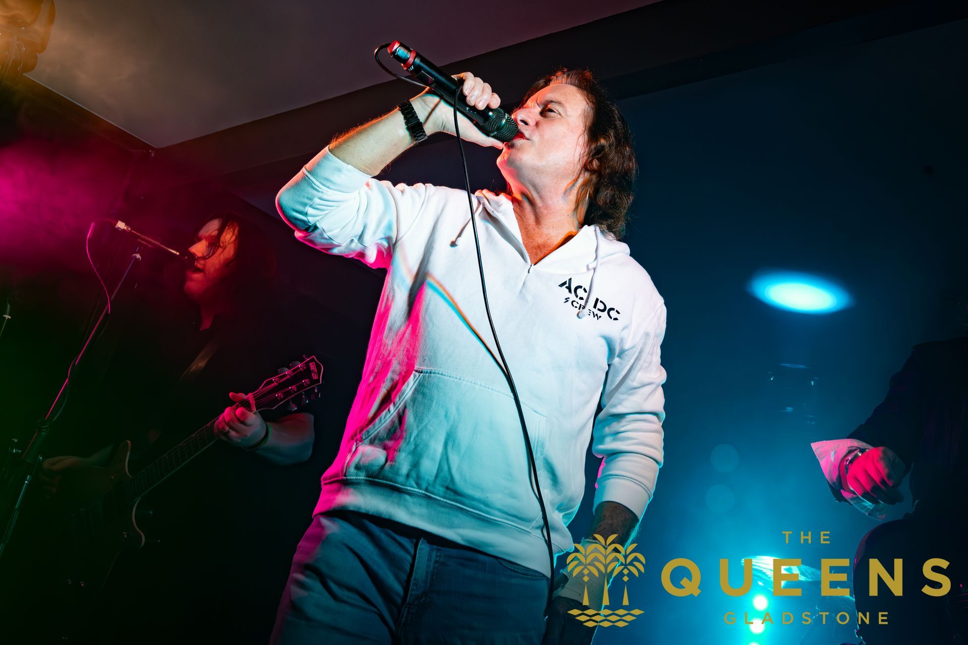 Singer performing onstage with a microphone, wearing a white hoodie under pink and blue stage lights.