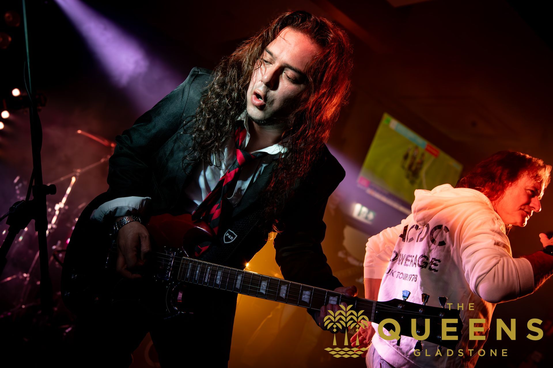 Guitarist onstage under red and purple lights, leaning into the performance with a black electric guitar.