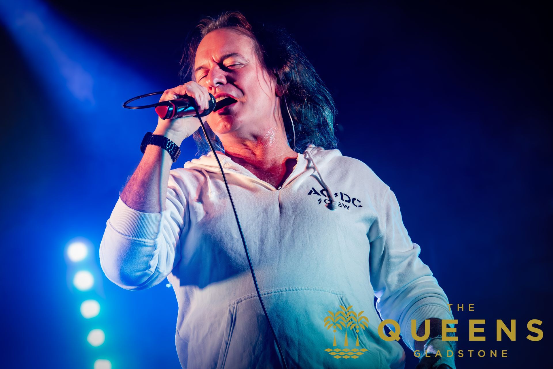 Singer performing on stage under blue lights, holding a microphone and wearing a white shirt.