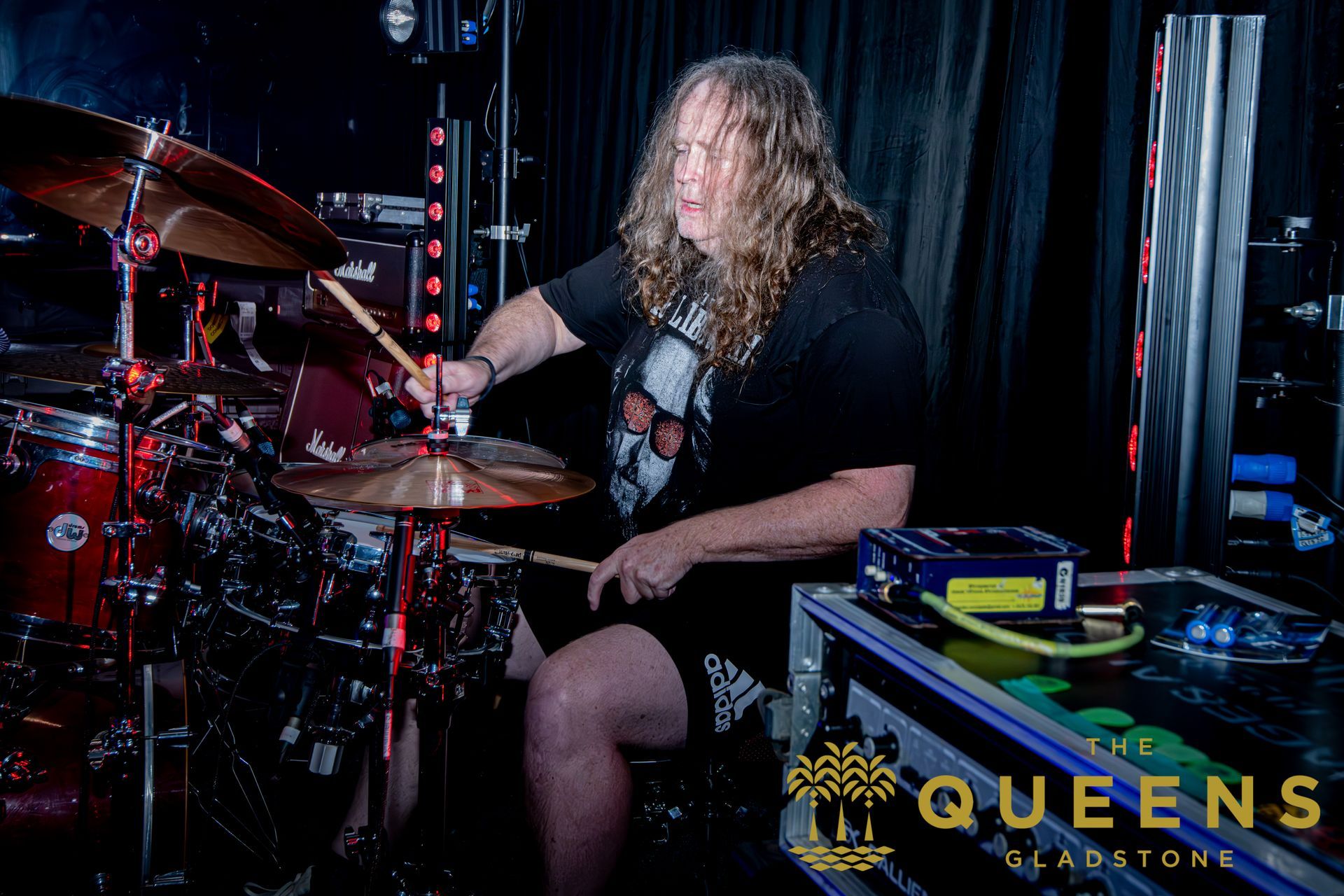 Drummer playing on a dark stage under red lights, with a “Queens” logo in the corner