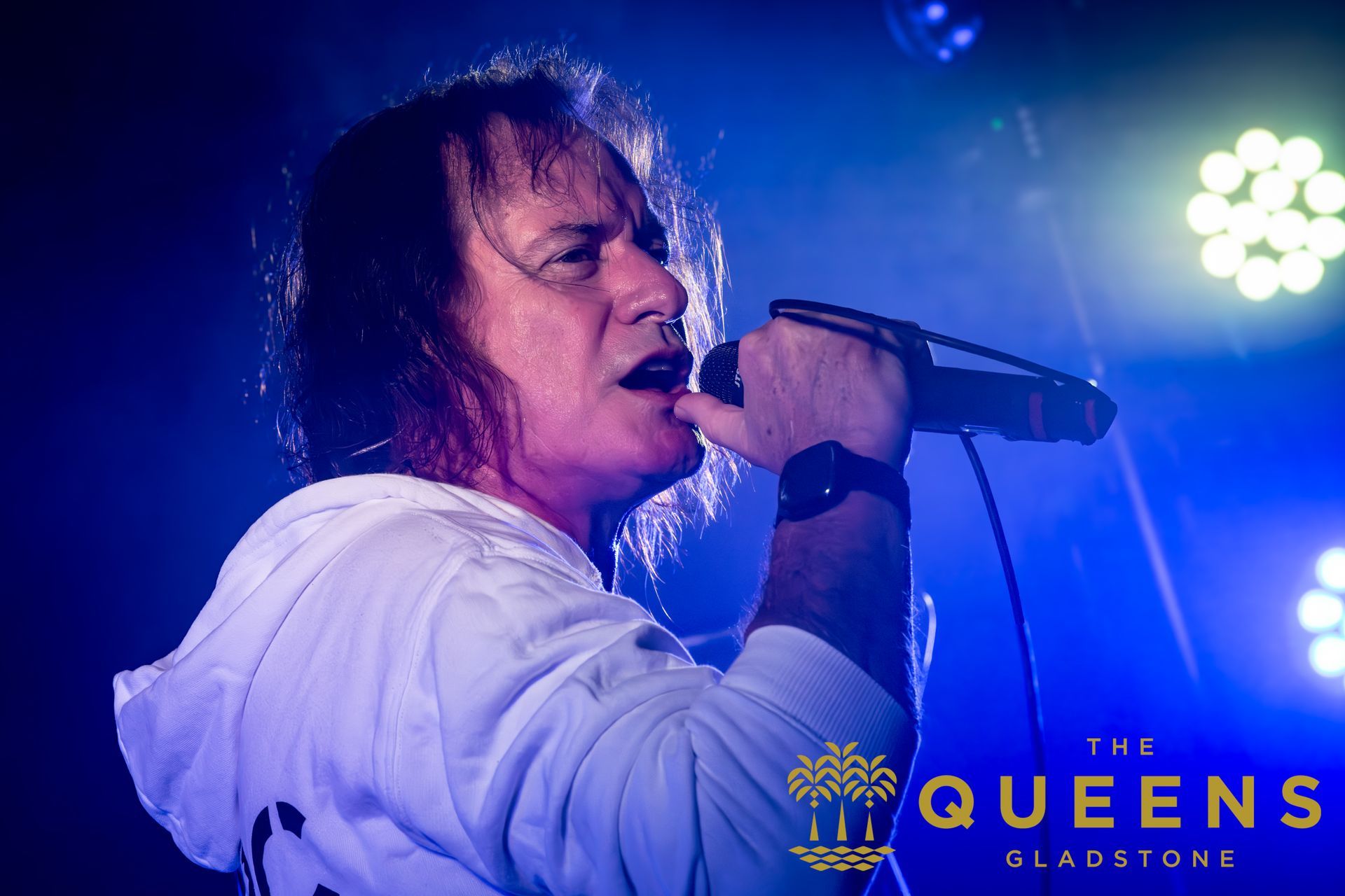 Singer in white shirt performing on blue-lit stage with microphone, The Queens logo at bottom right