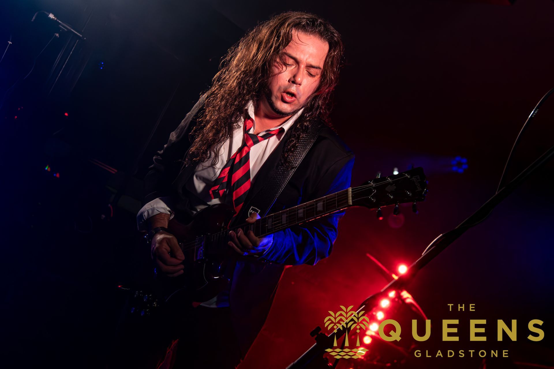 Guitarist performing onstage under red and blue lights with “The Queens” logo in the corner