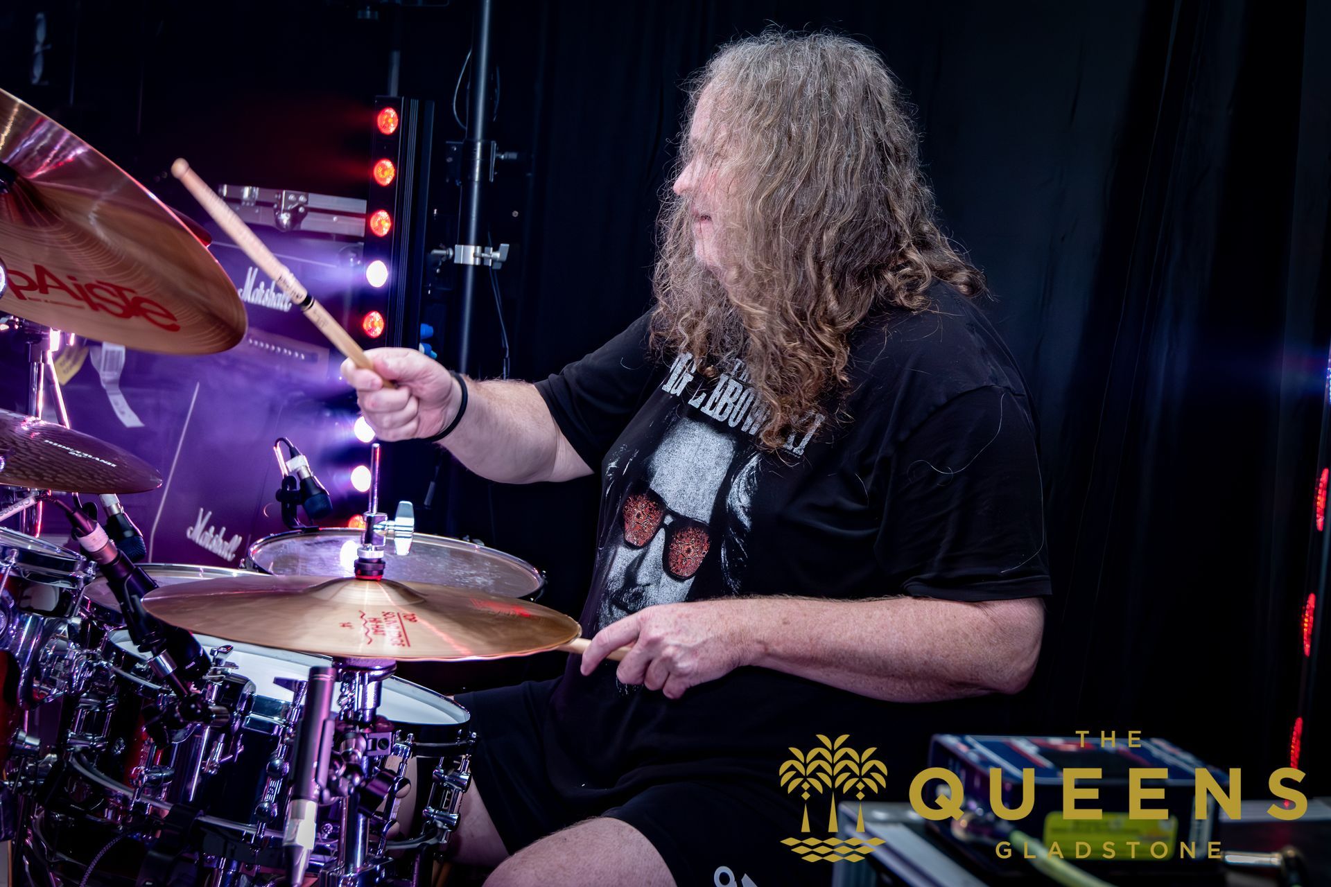 Drummer with curly hair playing on stage under purple lights, Queens logo in corner