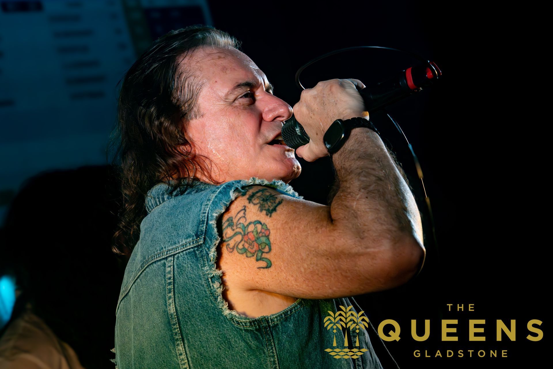 Singer in a denim vest performing into a microphone onstage, with “THE QUEENS” text in the corner