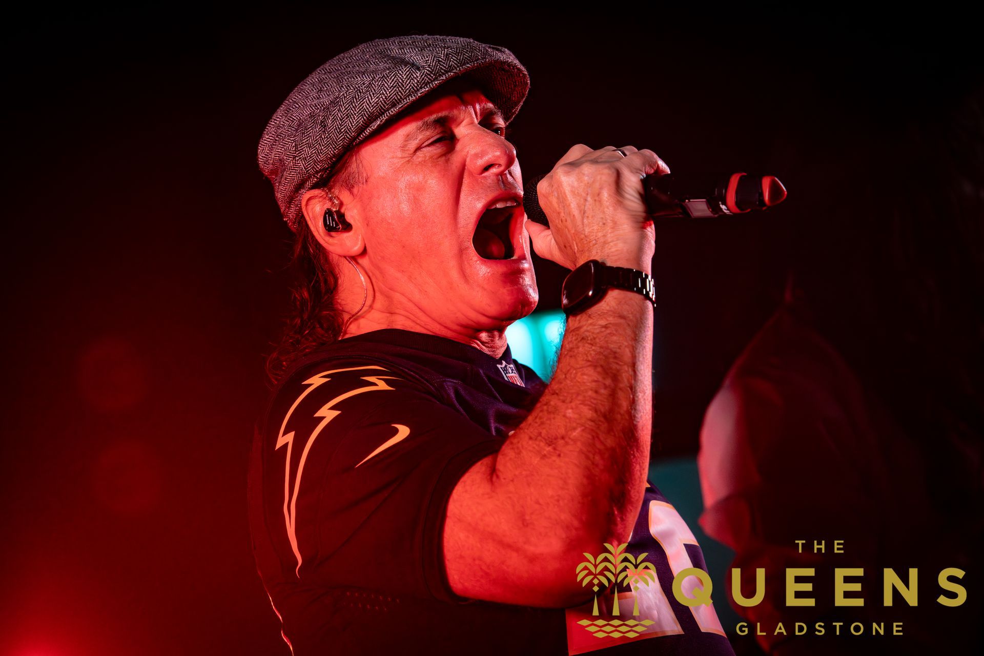 Singer in a flat cap performing on a red-lit stage with a microphone, mouth open mid-song