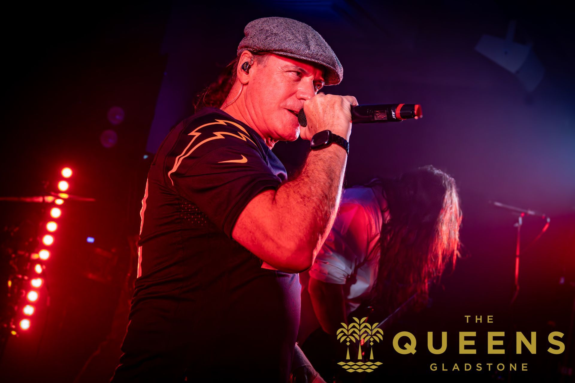 Singer performing on stage with microphone under red lights, with “The Queens Gladstone” logo