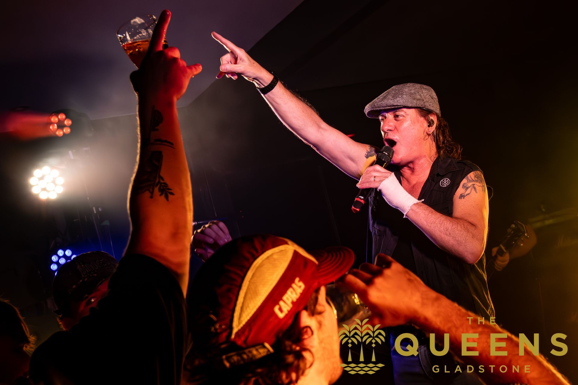 Singer on stage pointing to the crowd under red lights, with raised hands in the audience and “QUEENS” visible.