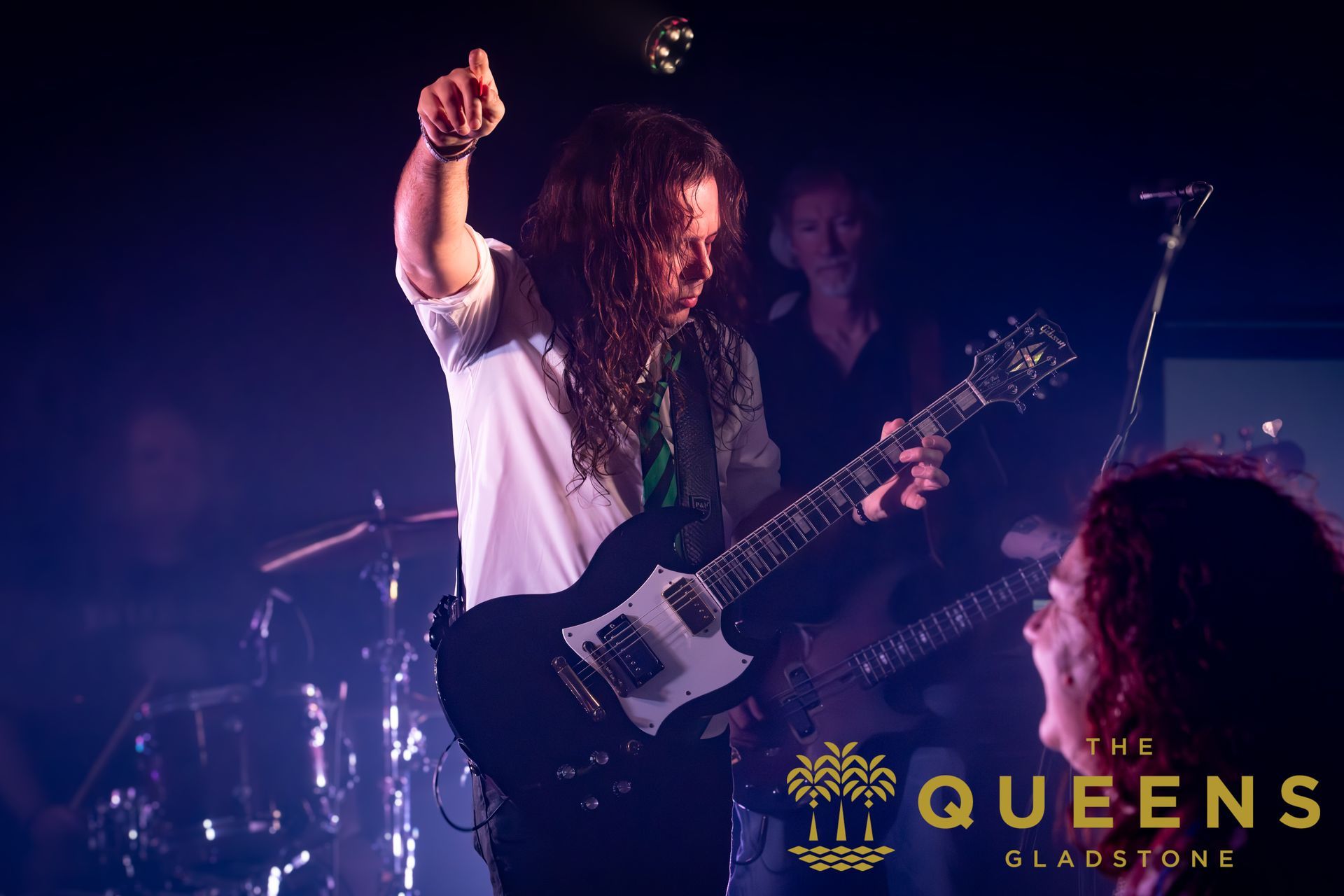 Rock guitarist onstage raising a fist under purple lights, with drums in the background and a QUEENS logo