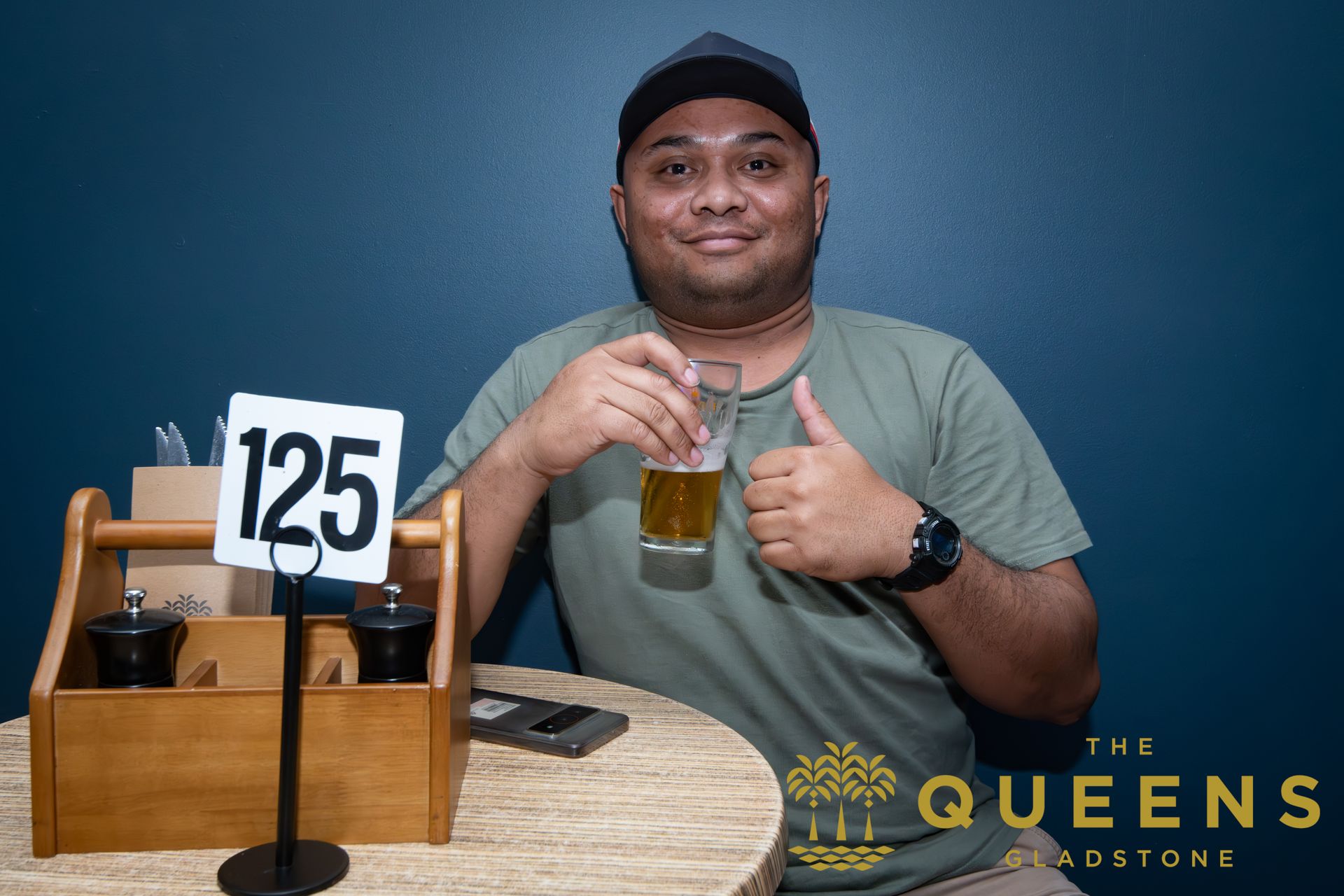 Man smiling at a table holding a drink beside a number 125 sign at The Queens restaurant