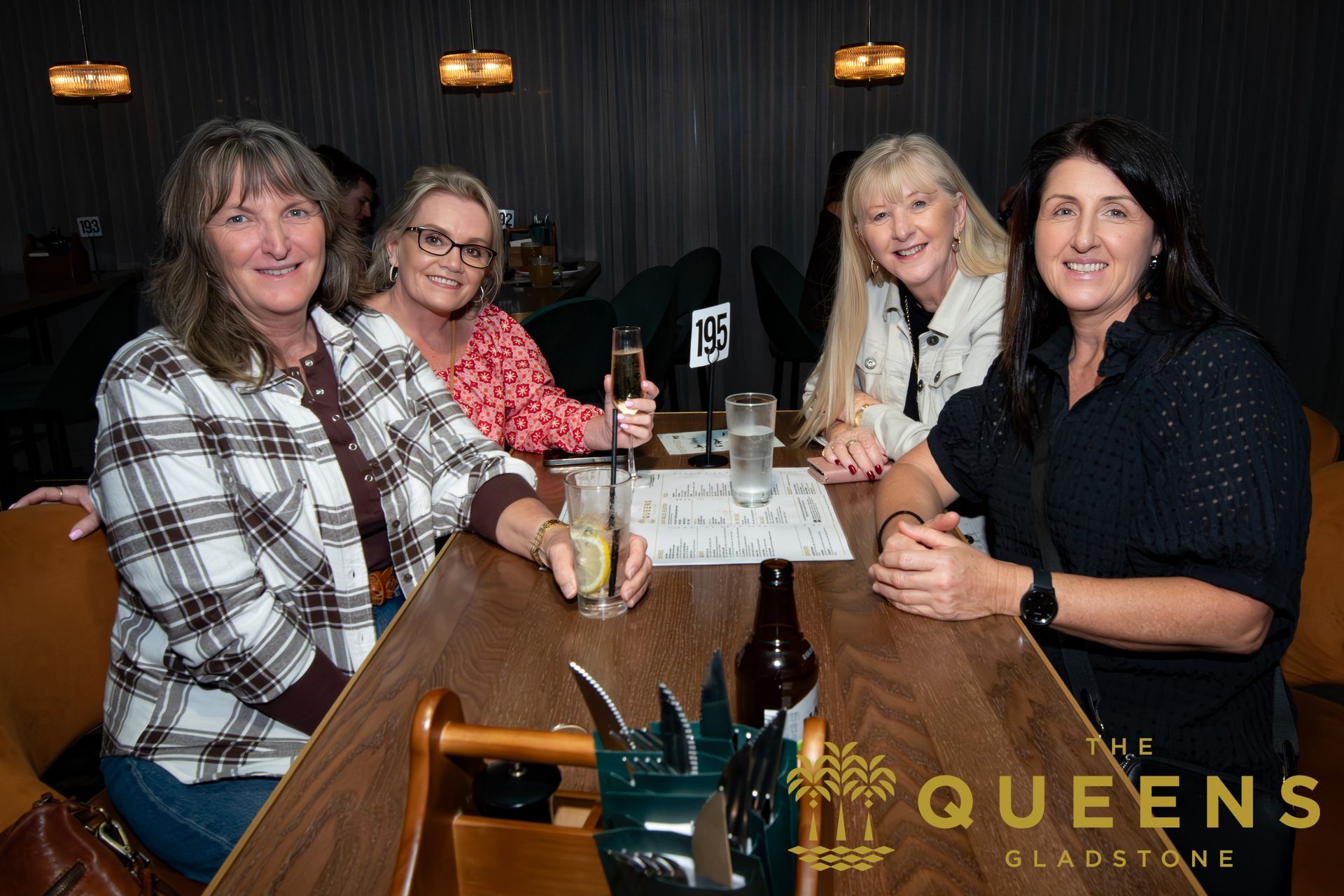 Four people seated at a restaurant table, smiling toward the camera, with drinks and a Queen’s-themed logo.