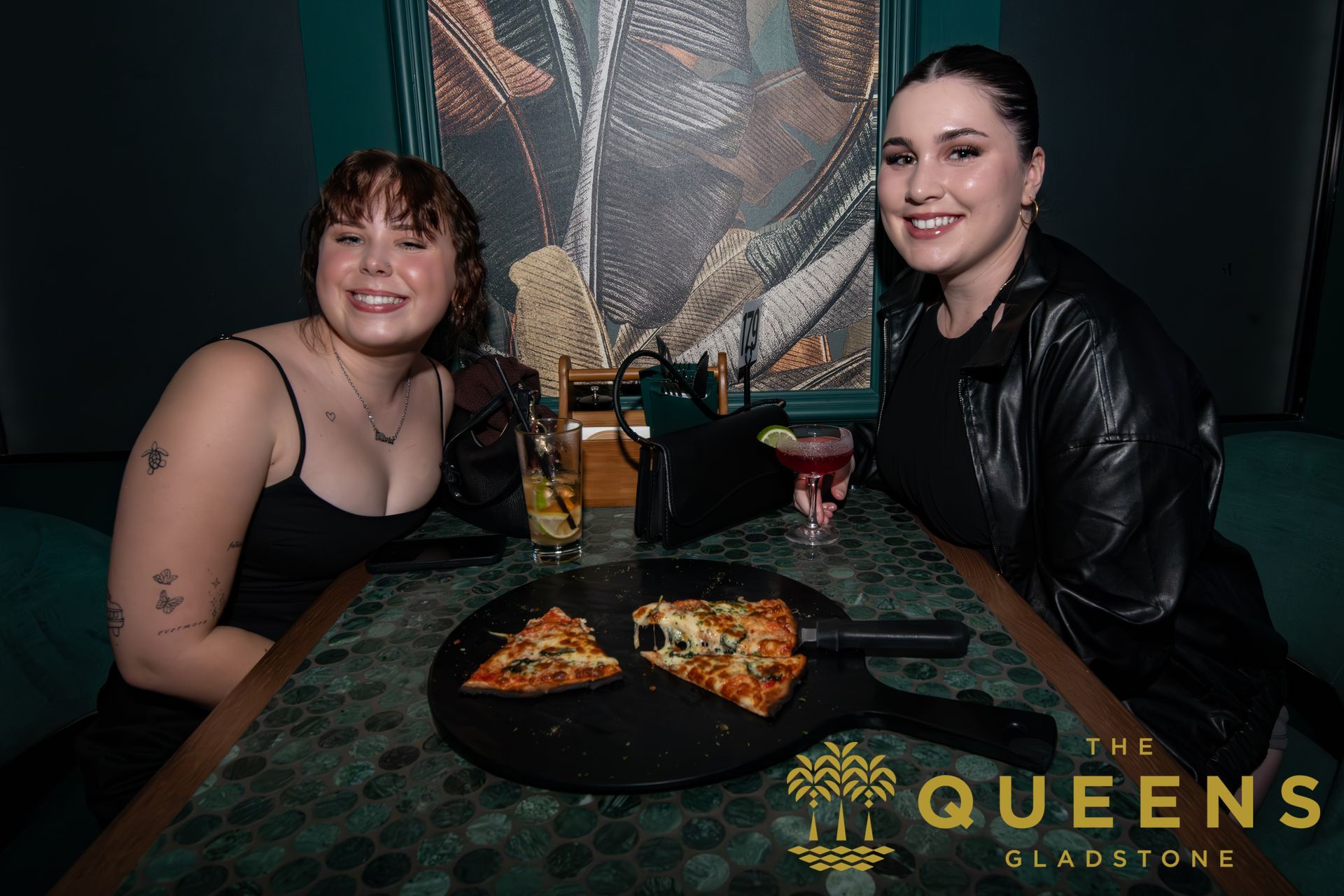 Two people smiling at a restaurant table with pizza and drinks, The Queens logo visible.