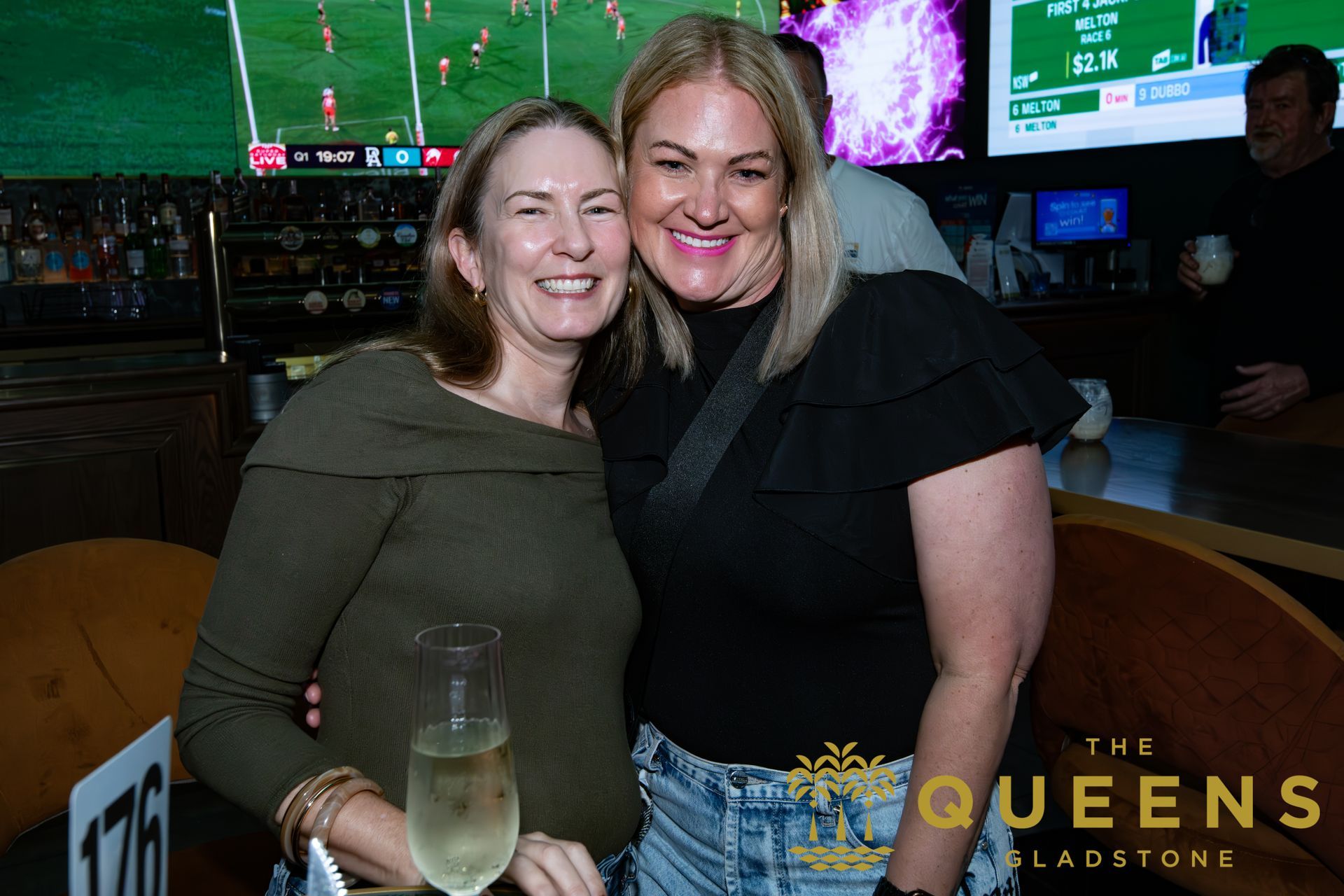 Two women smiling at a sports bar, with TVs in the background and a glass in front.