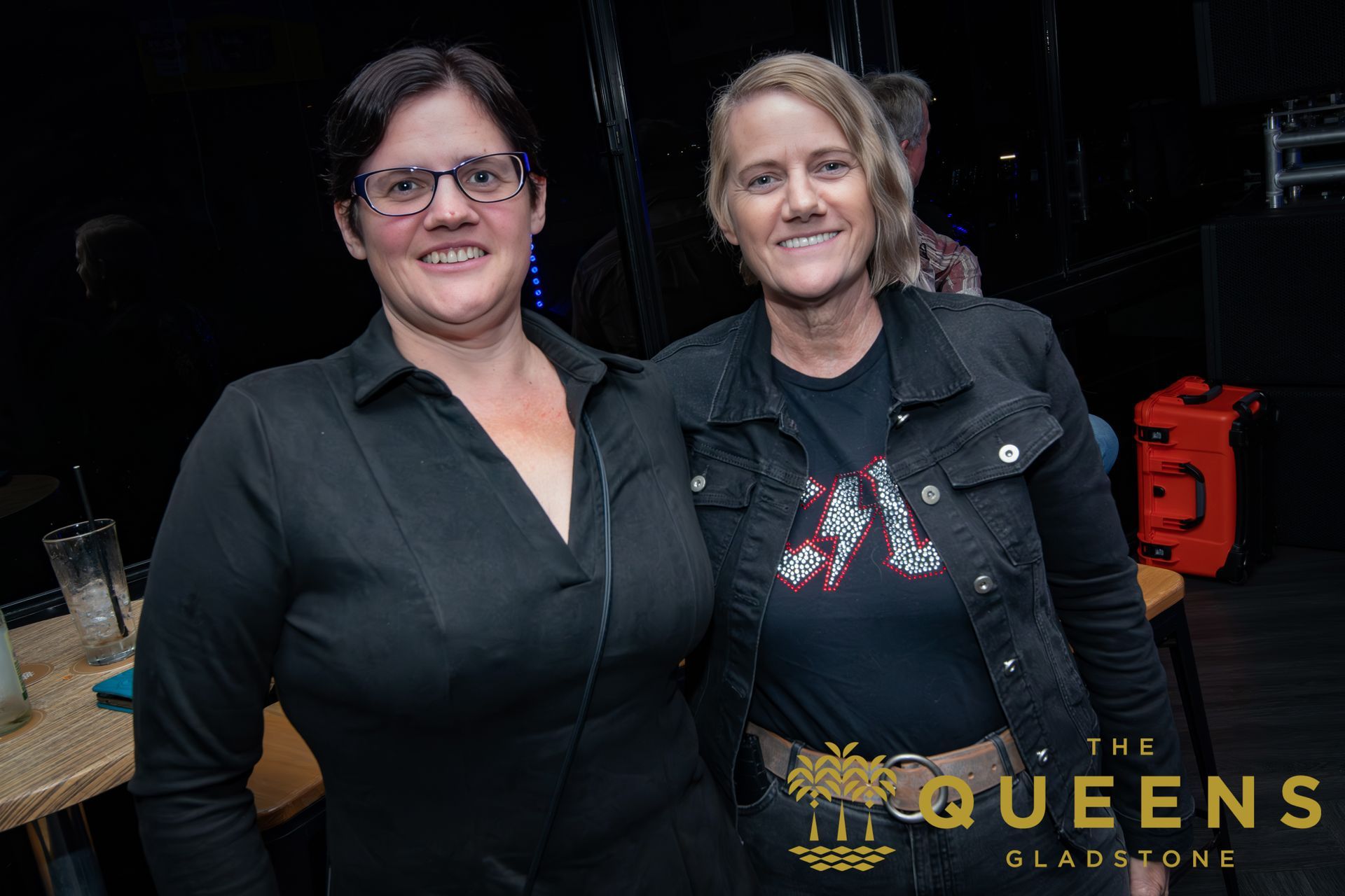 Two women posing together at a dark event backdrop, one in black and one in a black jacket with a graphic shirt