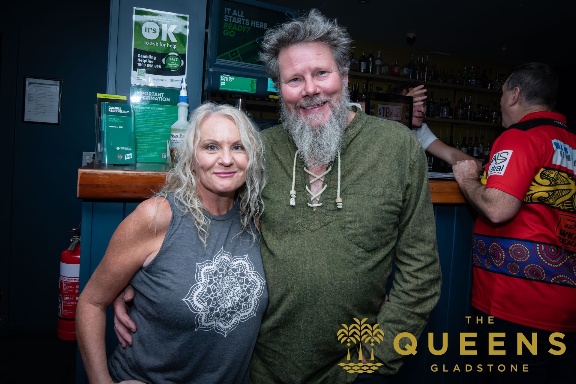Two smiling people pose in a bar, with a “The Queens” sign in the background.