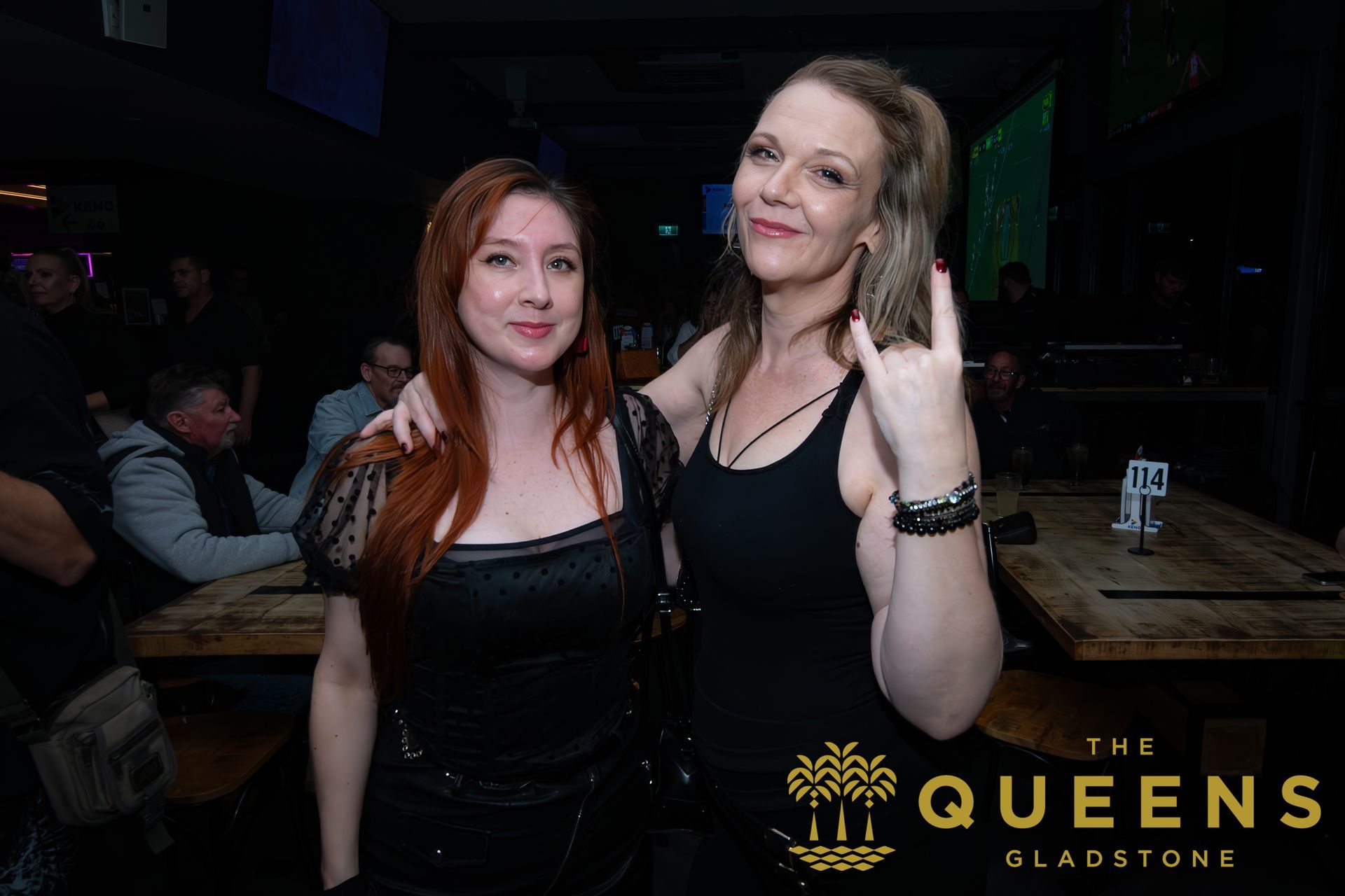 Two women posing in a dim nightclub; one flashes a peace sign, “The Queens Sladstice” logo at bottom right.