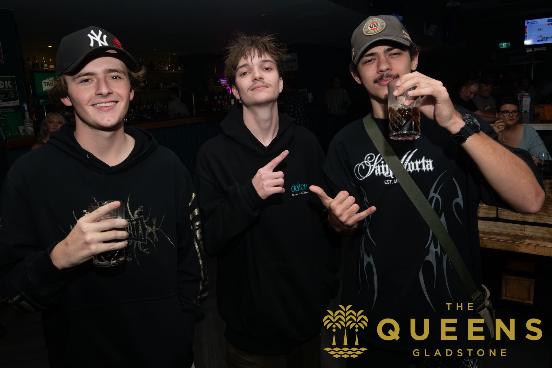 Three people posing with drinks at a dim bar, smiling and making hand gestures